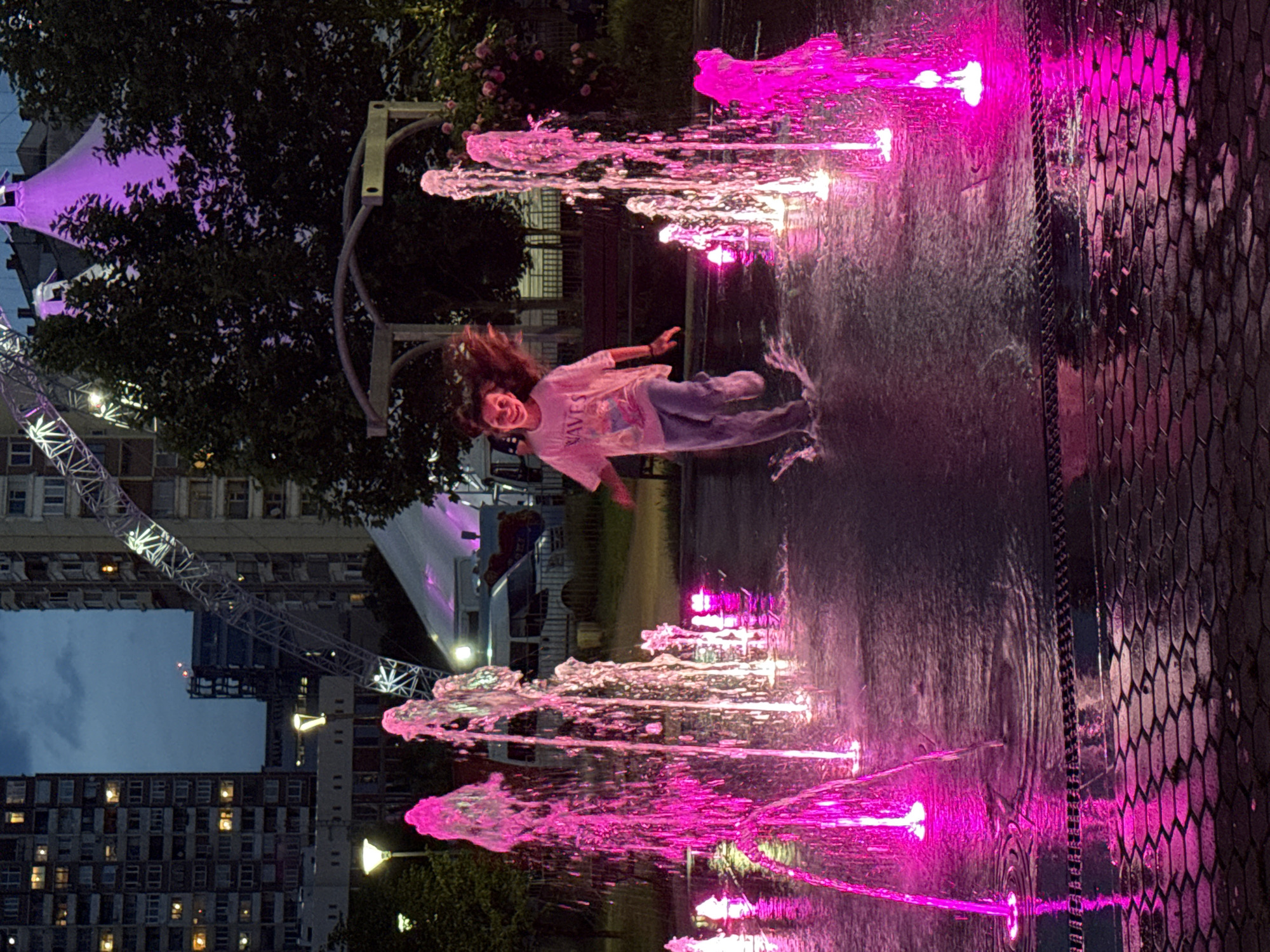 The image depicts an evening scene in a public park or festival area in Sofia, Bulgaria, likely at South Park during an event like Sofia Summer Fest. In the foreground, a young female with light brown hair, wearing a light-colored t-shirt with "WAVES" printed on it and light blue pants, runs joyfully through a water fountain. Her hair is in motion, and she is smiling directly at the viewer, creating splashes around her feet.
The fountain features numerous jets of water illuminated with vibrant pink and magenta lights, casting reflections on the wet, dark hexagonal pavement. In the mid-ground, trees are interspersed with urban elements. To the left, a streetlight with a warm yellow glow is visible. Towards the right, two distinct pointed tent structures, illuminated with purple light, stand behind trees. Near these tents, a white van and another vehicle are visible.
In the background, a large metal arch structure, consistent with a Ferris wheel or decorative arch, extends from the right, adorned with white lights. Tall residential buildings with various illuminated windows rise in the distant background, silhouetted against a deep blue evening sky. The overall scene is characterized by artificial lighting against the twilight sky.