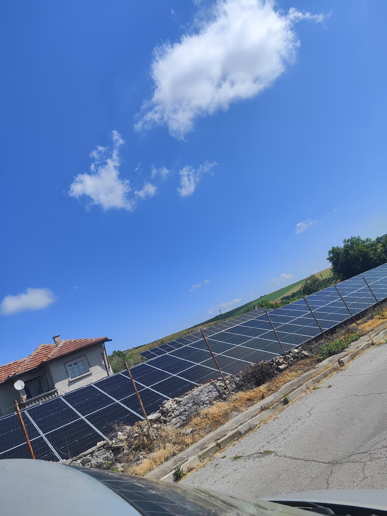 In Golyam Manastir, Bulgaria, a clear blue sky with scattered white clouds forms the backdrop for a residential structure and a solar panel array. The house, with a tiled roof, sits to the left, partially obscured by solar panels. The solar panels extend across the middle ground, arranged in neat rows and angled towards the sun. A paved road curves into the foreground on the right. The landscape beyond the solar panels consists of rolling green fields and trees, indicating a rural setting. The overall scene captures a sunny day in a Bulgarian village, with a focus on the integration of renewable energy technology.