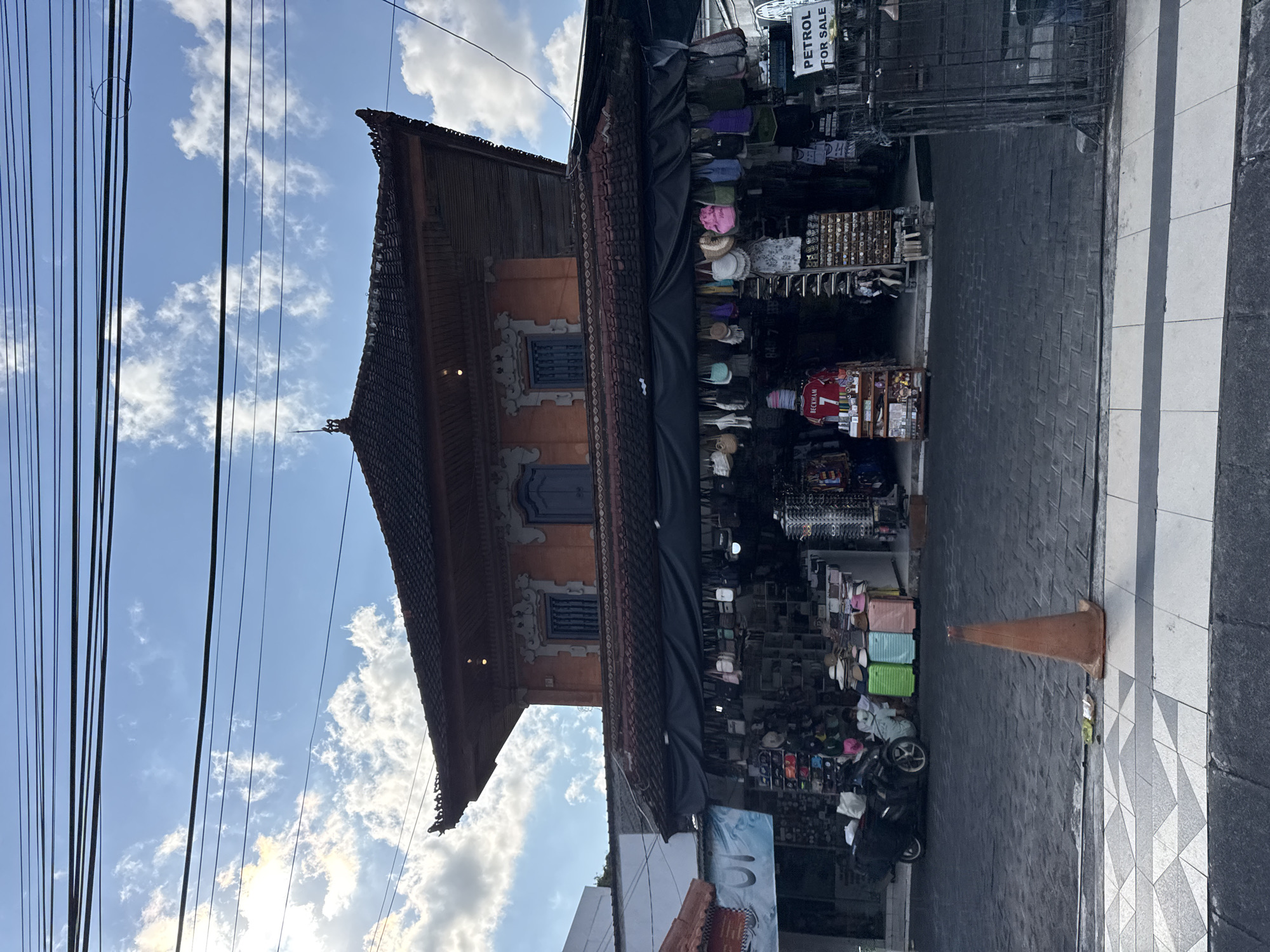 This vibrant street scene captures a sunny day in what appears to be a commercial area, consistent with the location context of Kuta, Indonesia.
On the left side of the image, a traditional Balinese-style building stands prominently. It features an ornate, multi-tiered dark brown roof with decorative eaves and orange-hued walls. Intricate carvings frame dark blue windows and adorn the building's facade. Numerous black power lines crisscross overhead against a clear blue sky dotted with scattered white cumulus clouds.
To the right, a lively market stall lines the street, packed with a wide variety of merchandise. Shelves are filled with items such as hats, brightly colored bags (including pink, light blue, and green), and other souvenirs. A red sports jersey with "BECKHAM" and the number "7" is clearly visible among the goods. Above the stall, a white sign clearly reads "PETROL FOR SALE."
The foreground features a dark paved street and a patterned sidewalk with black and white geometric tiles. An orange traffic cone is placed on the street near the market, and a dark-colored motorcycle or scooter is parked further down. The bright sunlight suggests it is daytime, likely morning or early afternoon, contributing to a bustling atmosphere typical of a tourist shopping district. No people are explicitly visible in the frame, but the open market implies commercial activity.