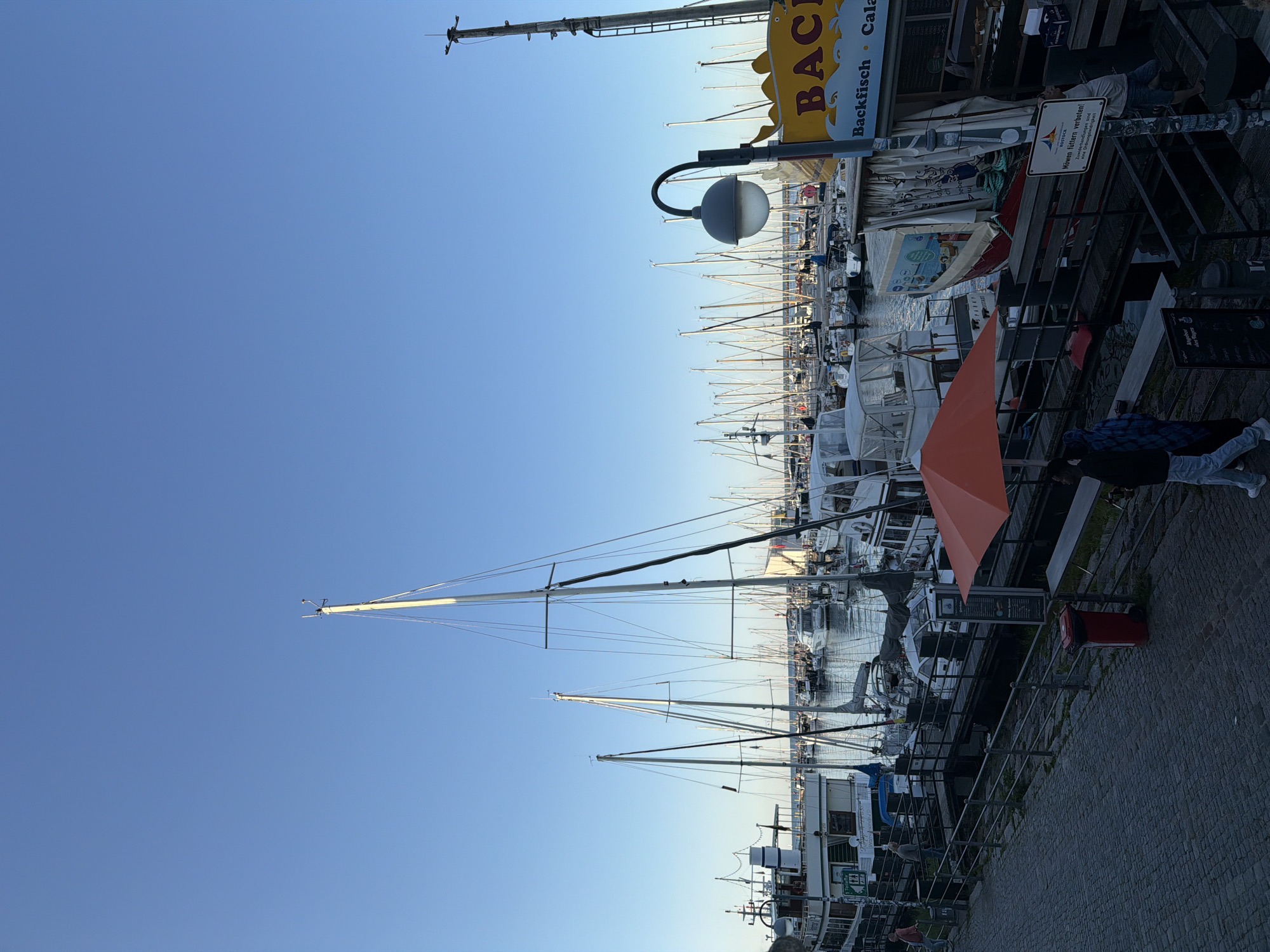 This image depicts a harbor scene in Rostock, Germany, likely in the early evening or late afternoon, given the clear sky and soft lighting. Several sailboats and a larger vessel are docked in the water, their masts creating a dense forest against the blue sky. A cobblestone pathway runs along the foreground, leading to a pier where people are seen walking and interacting.
On the left side of the image, a large white boat with dark trim is prominently docked, featuring multiple decks and windows. A green sign with text is visible on its side. Further down the pier, an orange umbrella is set up, casting a shadow. A red trash bin is positioned near the pathway.
To the right, a yellow sign with blue lettering that appears to be a food vendor is visible, with text that includes "BACKFISCH" and "CALAMARI." A street lamp with a grey spherical shade curves over the pathway. People are scattered throughout the scene, some walking along the pier and others possibly near the boats. The overall atmosphere suggests a calm and picturesque maritime setting in Rostock.