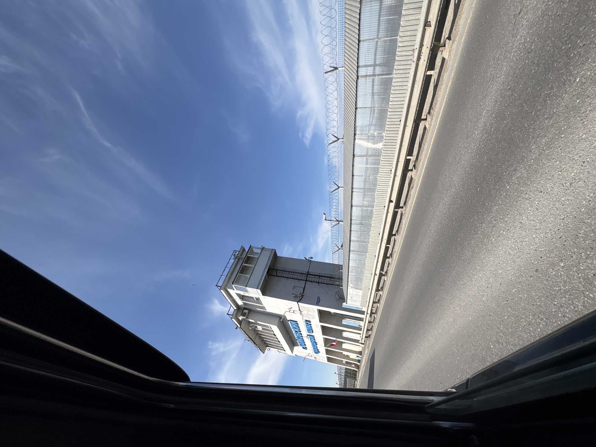 The image shows a view from inside a car through an open sunroof, with a partial view of the car's interior. The background features a tall, multi-story white building with blue signage written in Russian, located on a road in the city of Chaikovsky, Russia. There appears to be a barrier along the road with razor wire on top. The sky is blue with light clouds. The road surface is gray.