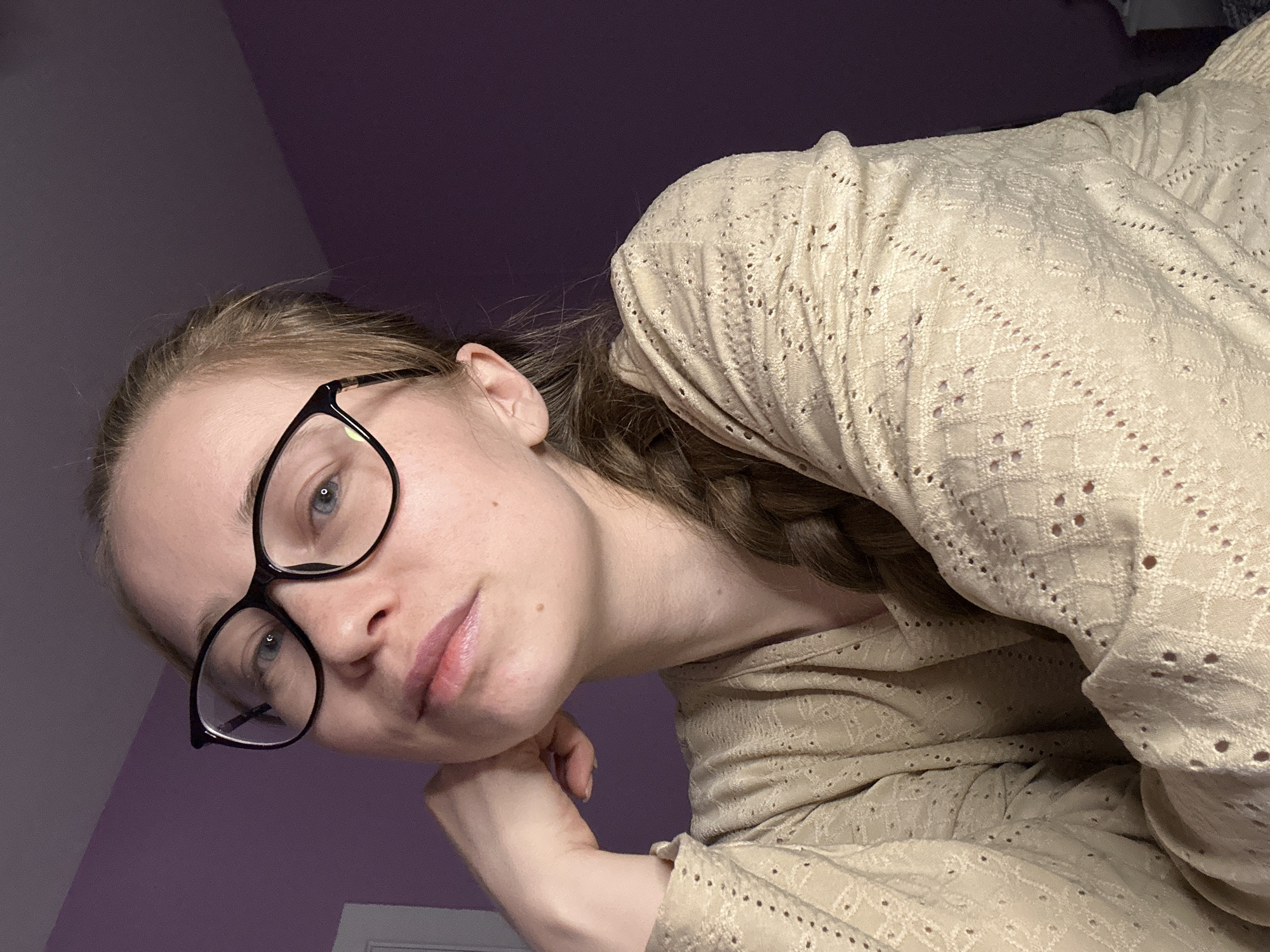 A fair-skinned young adult female with light brown hair is leaning towards the camera, her cheek resting on her left hand. She is wearing black-rimmed glasses and a light beige, long-sleeved, patterned shirt. The background is a plain, light purple wall. The scene provides no information to suggest it is located in Saint Petersburg, Russia.