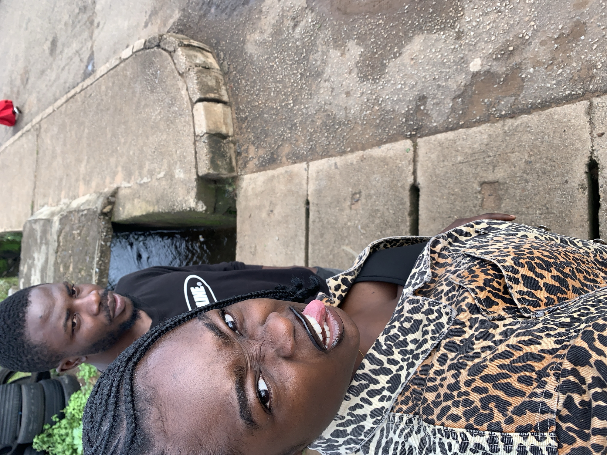 In the city of Rafin Par, Nigeria, two individuals are captured in a portrait-style photograph. The foreground is dominated by a young woman with braided hair, her face angled towards the camera with an exaggerated expression, sticking out her tongue and scrunching her nose. She is wearing a black top underneath a leopard-print jacket. Behind her, a man with short hair and a beard, wearing a black t-shirt with a white logo, looks slightly off-camera.
The setting appears to be outdoors, with a concrete pathway and a narrow, dark-colored channel or drain to the left. In the background, a stack of black tires and some greenery are visible. Further to the right, the concrete path continues, with a person in a bright red garment walking away from the camera in the distance. The ground surface is a mix of concrete and what appears to be a gritty, possibly wet, pathway. The lighting suggests an overcast day.