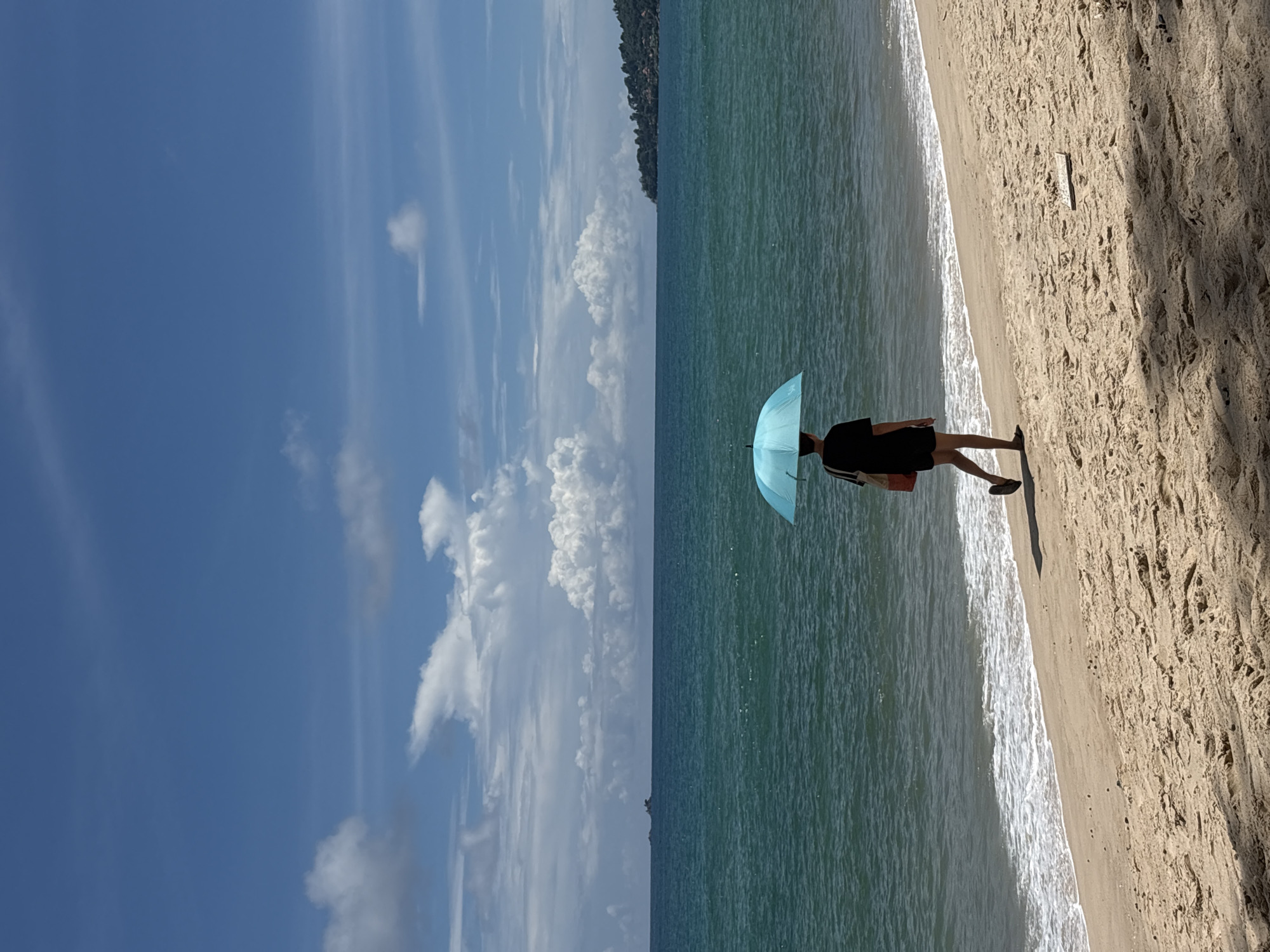 A lone person walks along a sandy beach in Choeng Thale, Thailand, holding a light blue umbrella to shield themselves from the sun. The person, wearing a black t-shirt and black shorts, is carrying a light-colored bag over their shoulder. The beach has a gentle slope towards the turquoise ocean, where small waves are breaking. In the distance, the coastline is visible with lush green trees and some buildings, and a small island is visible further out to sea. The sky is bright blue with scattered white clouds. The sand on the beach is a light brown color with some textures and shadows from unseen objects.