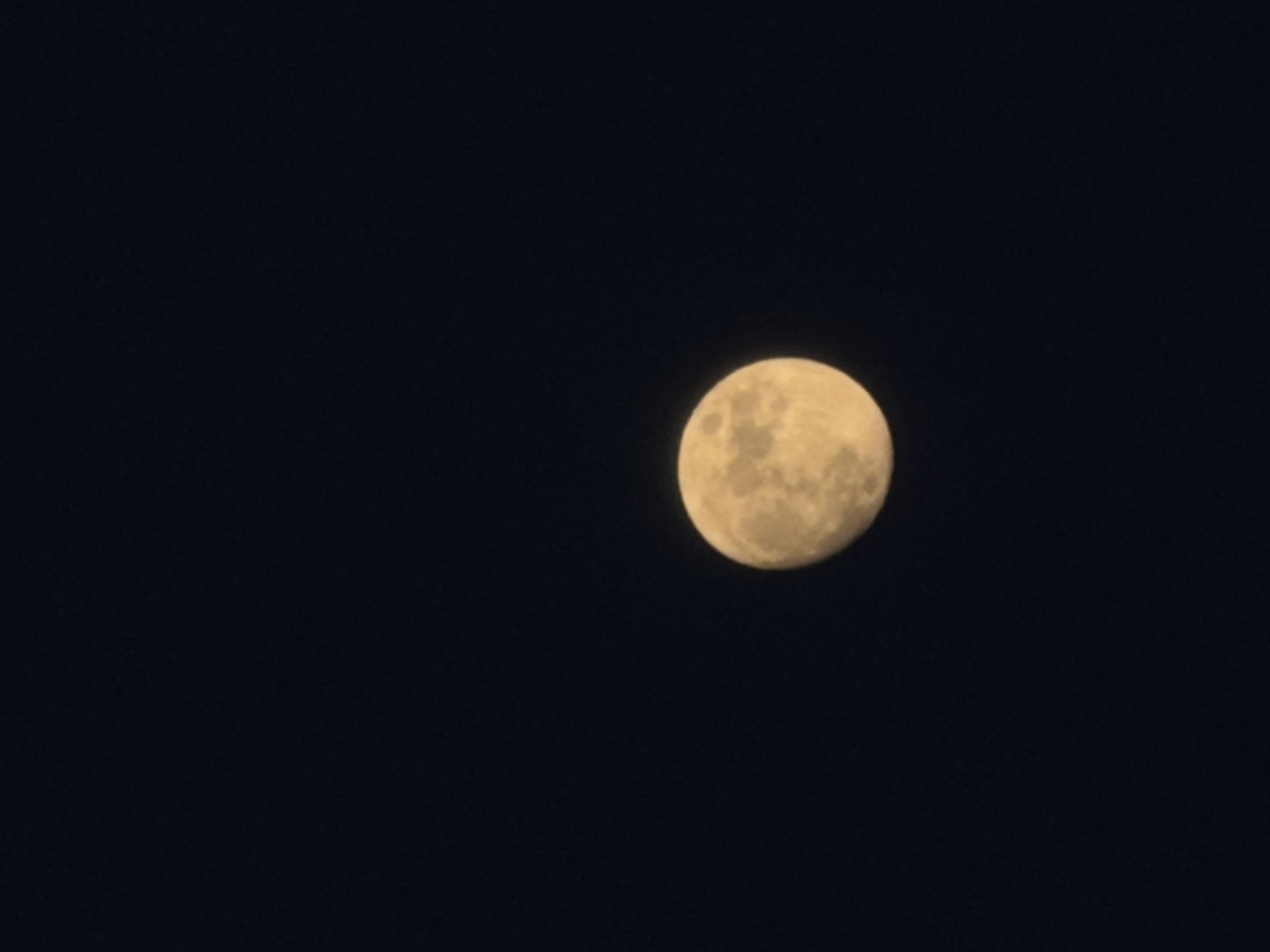 The image captures a bright, full or gibbous moon set against a dark, clear night sky. The moon, the primary subject, appears yellowish-white with discernible surface textures, including variations in brightness that suggest craters and maria. It is positioned slightly to the right of the center of the frame. The background is a uniform, deep black, indicating a cloudless night and the absence of any other visible celestial objects. The scene provides a focused, tranquil view of the moon during nighttime in Chaikovsky, Russia. There is no visible text or human activity.
