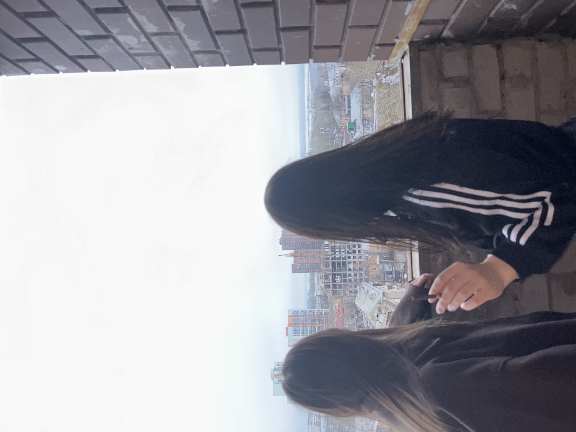 The image shows two people standing on the unfinished balcony of a high-rise building. The building appears to be under construction in Perm, Russia. The viewpoint overlooks a cityscape in overcast weather. One person is wearing a black jacket with white stripes on the sleeves. The brickwork of the building surrounds the view of the city.