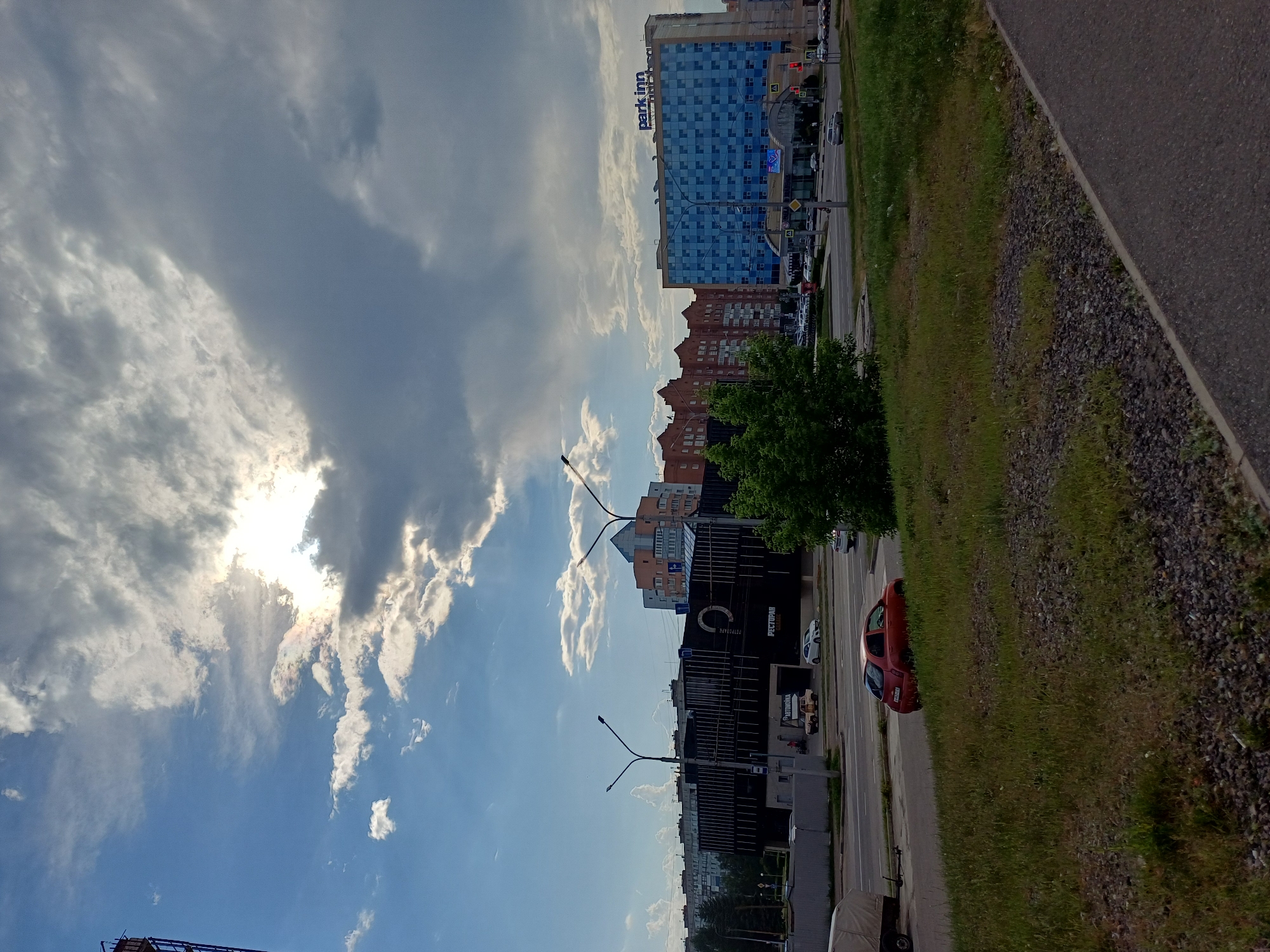 The image depicts a cloudy sky over a city street with buildings and vehicles. In the foreground, a grassy embankment with gravel is visible, adjacent to a paved path. A red car is parked on the right side of the street. Further down the street, a dark building with vertical lines is visible, with a sign that appears to say "РЕСТОРАН" and "АВТОМОЙКА". Another building, blue with a checkered facade, has the text "park inn" on its rooftop. Several other buildings and trees are also present in the scene, along with streetlights and traffic signals. The sky is partially obscured by large, grey clouds, with patches of blue sky and sunlight breaking through in the upper left portion of the image.