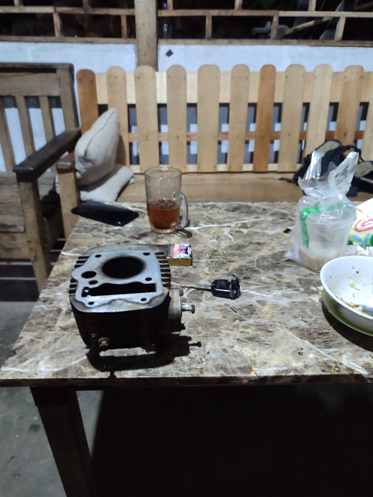 In Gantungan, Indonesia, on a table with a marble-patterned top, lies a disassembled motorcycle engine cylinder. Beside it, a cigarette pack and a key are visible. A glass of amber liquid, possibly tea, sits to the left, and a plastic bag containing an item is to the right. In the background, a wooden chair with a cushion is present, along with a light-colored wooden fence. The scene appears to be an outdoor or semi-outdoor setting, possibly a yard or patio, with dim lighting.