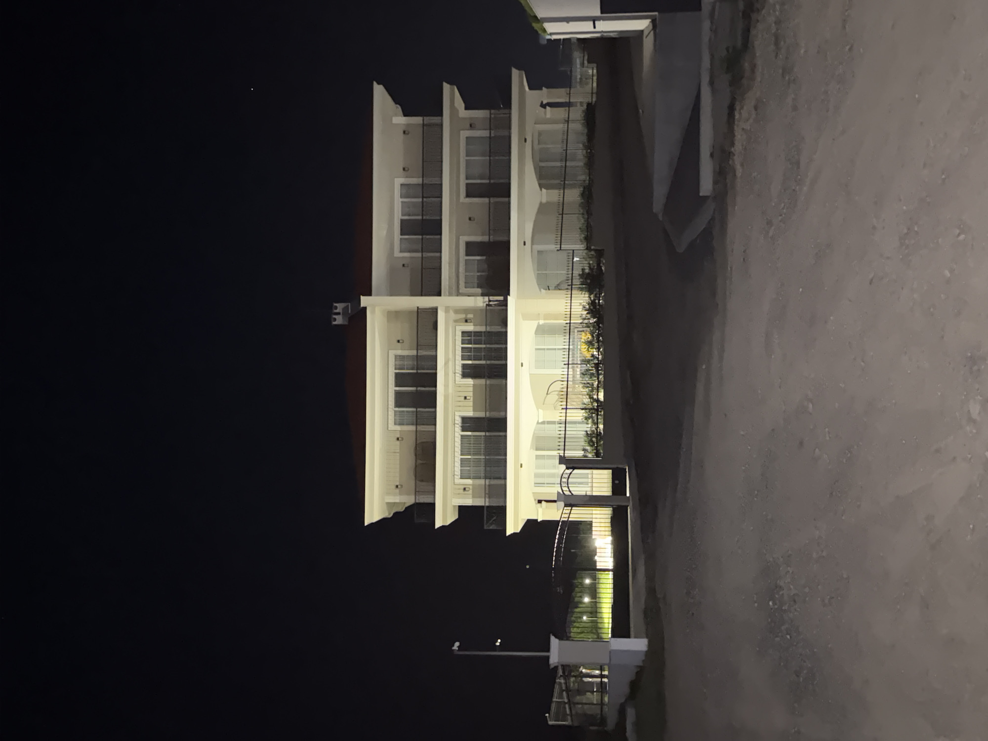 The image depicts a modern, multi-story building at night, likely in the city of Chernomorets, Bulgaria, given the context. The building is illuminated by artificial lights, highlighting its cream-colored facade and dark balconies. It features multiple levels with numerous windows and balconies, suggesting it could be an apartment complex or a large residence. A red-tiled roof caps the structure.
In the foreground, a dirt or gravel path leads towards the building. To the left, a dark metal gate with vertical bars is visible, partially enclosing an area with lights illuminating what appears to be a lawn or garden. A lamppost stands near the gate. The overall environment suggests a suburban or semi-rural setting, given the unpaved road and the presence of green space. The sky is dark and clear, with a few faint lights visible, possibly stars or distant lights. No people are visible in the image, and the scene appears quiet and still.