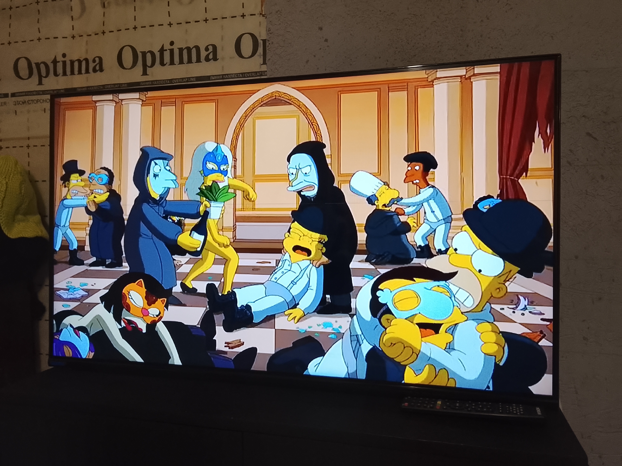 The image shows a television screen displaying a scene from an animated show that appears to be a parody or a reimagining of the film "Eyes Wide Shut," but with characters resembling those from "The Simpsons." The background features a luxurious interior with golden pillars and arches. Several characters are dressed in elaborate, costume-like attire. Some characters are depicted engaging in physical interactions, such as being struck or restrained. One character is lying on the floor. The environment includes broken glass and debris scattered across the floor. The television is placed on a black stand, with a remote control visible. The location, Высокиничи, Russia, is extraneous as it is not depicted in the image.