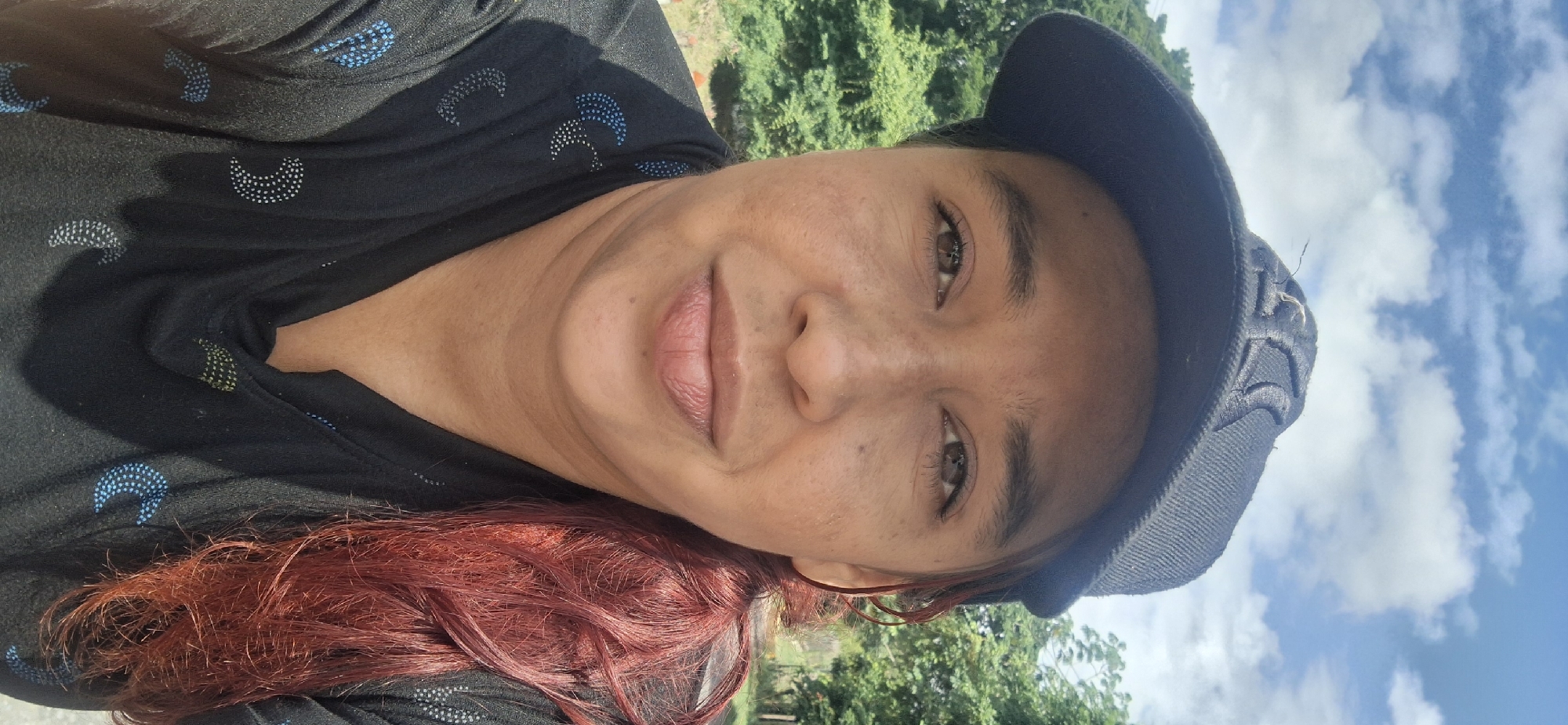 The image shows a person with medium brown skin, red-tinted hair, and brown eyes, wearing a black hat and a black top with crescent-shaped patterns. The person is positioned against a backdrop of green foliage and a blue sky with white clouds, suggesting an outdoor setting. While the specific location cannot be verified as Vallecito, Venezuela from the image alone, the vegetation is consistent with a tropical or subtropical environment.