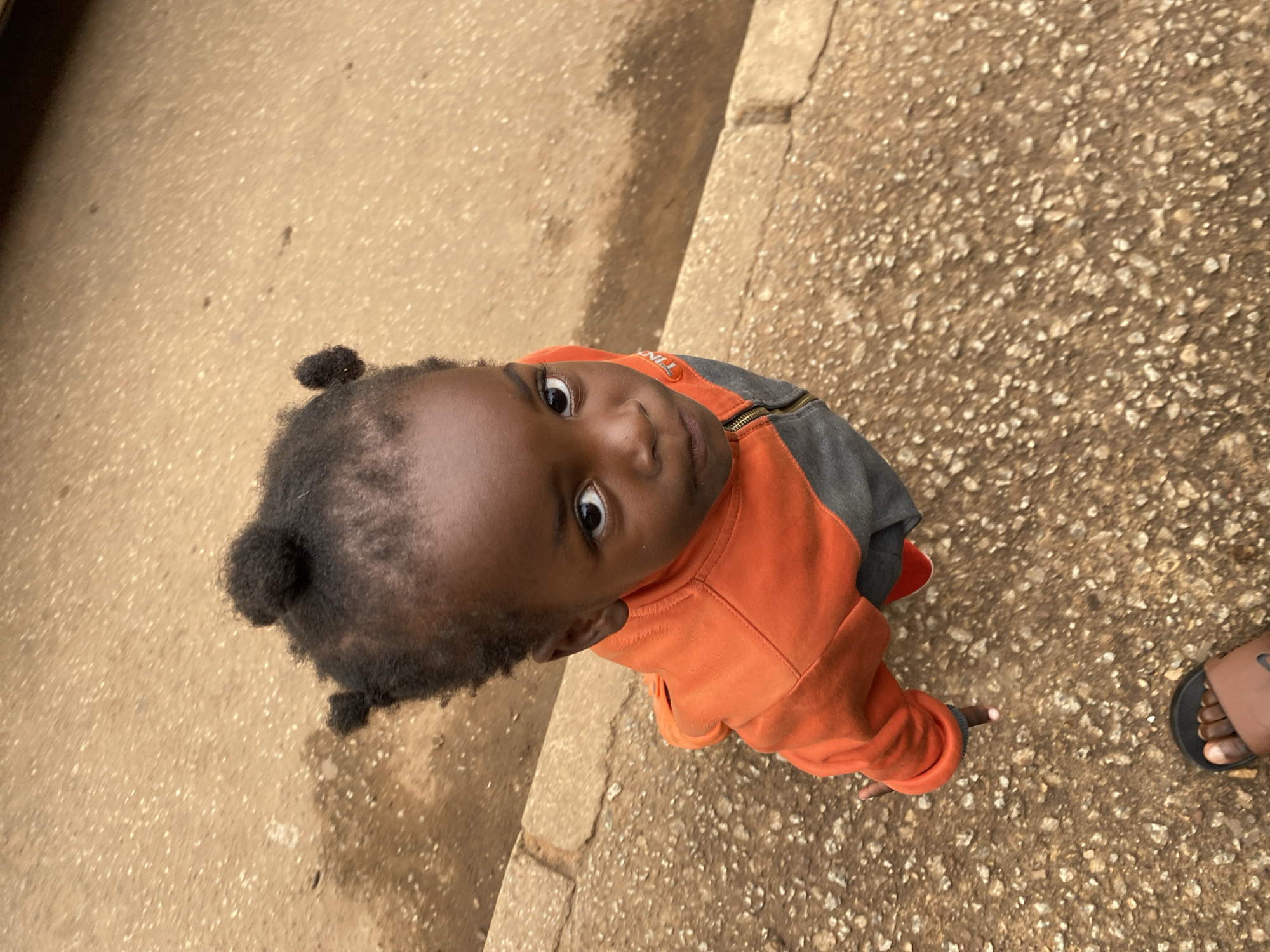 A close-up, high-angle shot captures a young child standing on a gritty, tan-colored paved surface in Bukuru, Nigeria. The child, facing directly towards the camera, has dark skin and hair styled into four small buns. They are wearing an orange and gray zip-up jacket. A hint of a red shoe is visible on the child's left foot. The ground beneath is composed of a mixture of dirt and small stones, with a concrete curb running along the left side of the frame. A person's bare foot, wearing a brown sandal, is visible at the bottom of the image. The overall lighting suggests an overcast day.