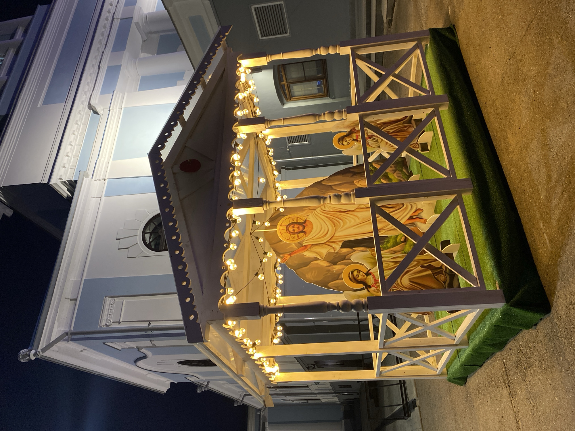 A low-angle shot depicts a white wooden nativity scene structure decorated with strings of lights, situated next to a light blue and white building. The nativity scene features painted depictions of religious figures. The structure rests on a patch of green material resembling grass. The photograph appears to be taken at dusk or night. Based on the presence of the orthodox church and the style of the decorations, this is likely located in Votkinsk, Russia.