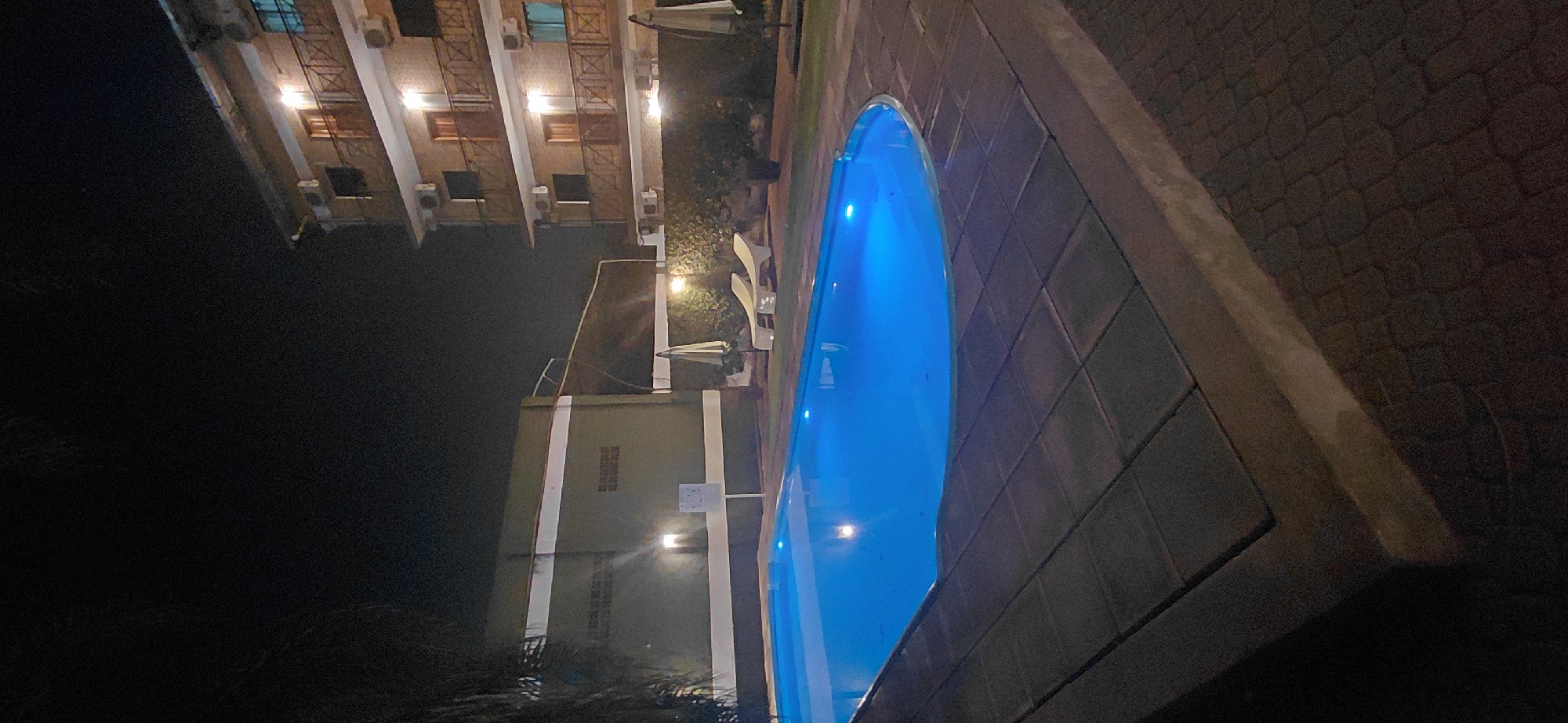 An exterior nighttime shot features a swimming pool illuminated by blue lights, with a building and other amenities in the background. The pool is rectangular with rounded corners and is surrounded by a tiled patio. To the right of the pool, a multi-story building with balconies and windows is visible, lit by interior lights. In front of the building, two lounge chairs and a large umbrella are set up on a grassy area. To the left of the pool, a dark, low-lying structure with lights is present. The overall scene suggests a hotel or resort setting in Presidente Franco, Paraguay.