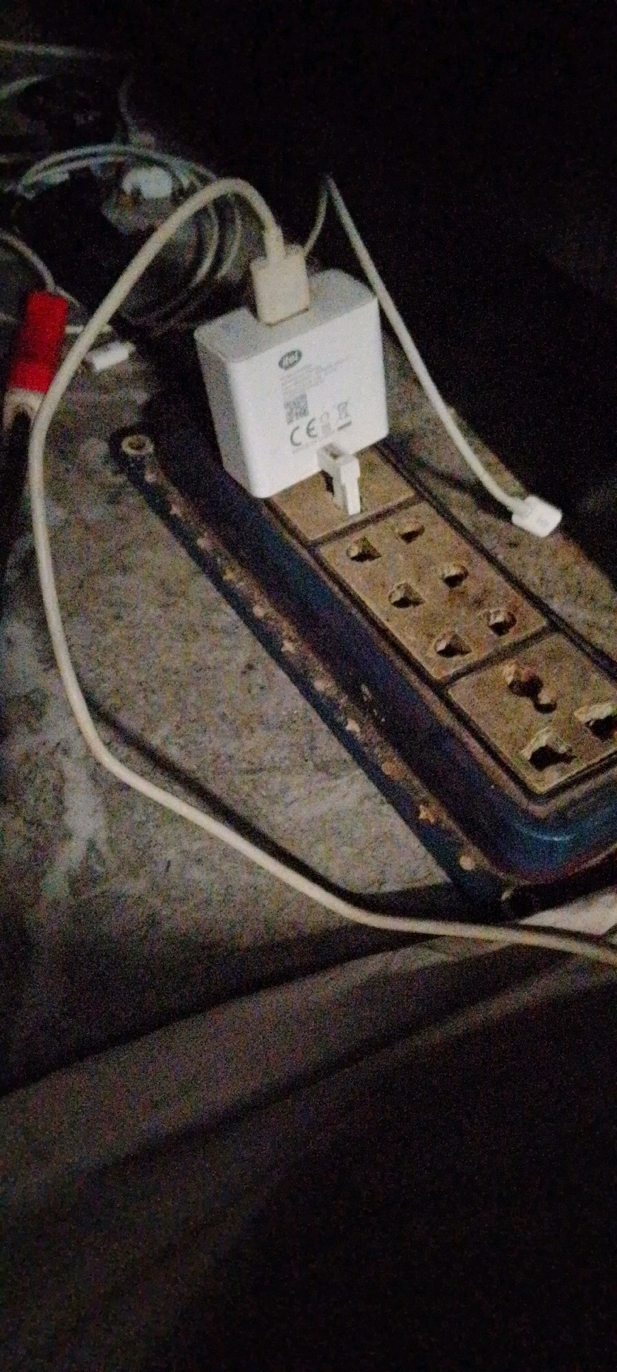 The image shows a close-up view of a power strip with multiple outlets, some of which appear to be occupied by plugs. A white wall adapter, labeled with "CE" and a QR code, is plugged into one of the outlets. A white cable extends from the adapter and appears to be connected to a device not fully visible in the frame. Another red object, possibly another cable or adapter, is partially visible to the left. The power strip is placed on a rough, textured surface, possibly a floor or a table. The background is dark and out of focus, suggesting an indoor environment. The overall impression is of a functional electrical setup, potentially in a home or business. The city of Oshodi, Nigeria, is not directly depicted or implied by the visual elements in the image.