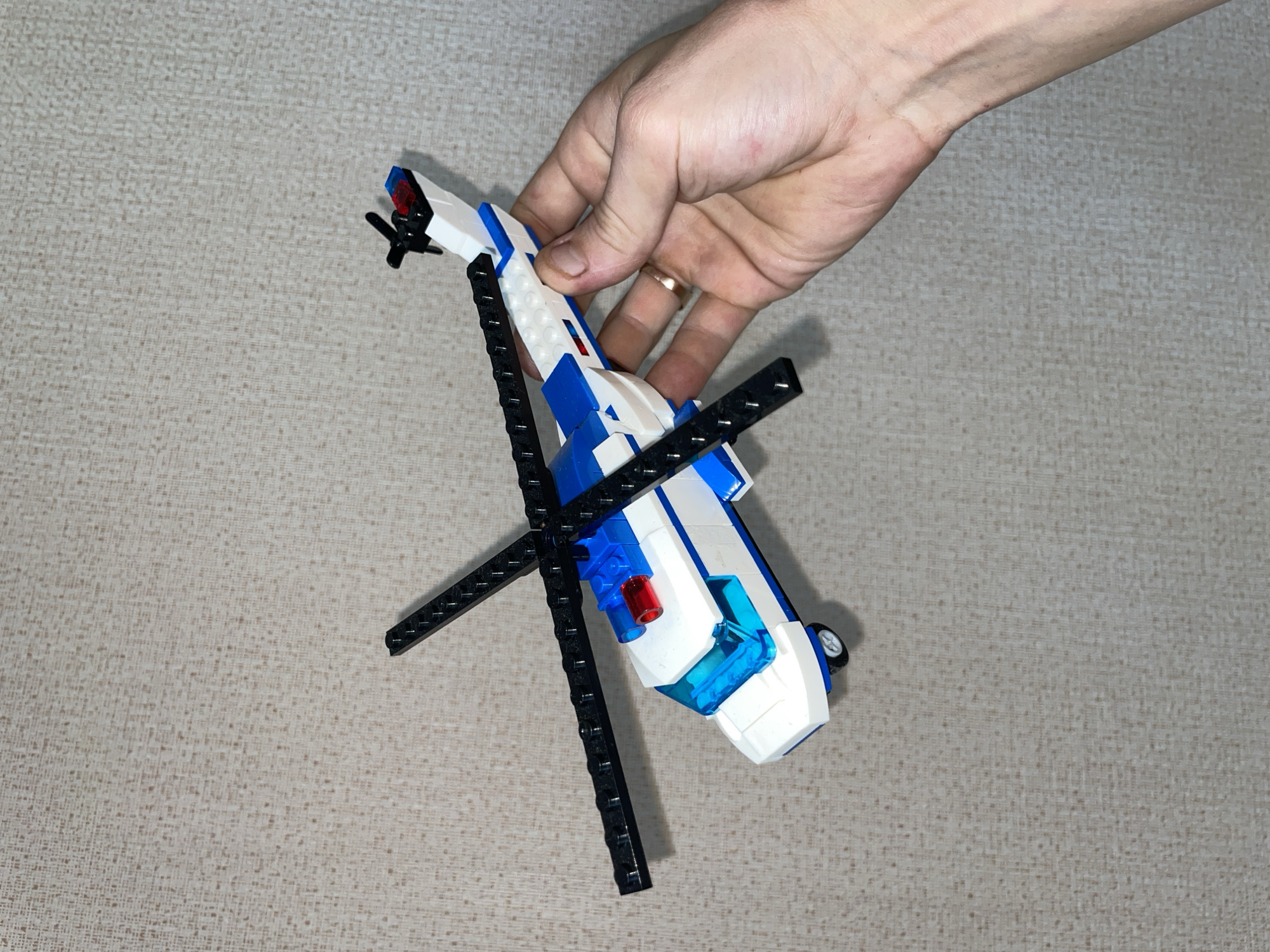 A Lego helicopter is held by a person's hand against a textured, light-colored background. The helicopter is primarily white and blue, with black rotor blades. A red and blue flashing light is visible on top of the helicopter's cabin, and a transparent blue windshield is present. The background appears to be a wall or fabric with a subtle, repeating pattern. The person's hand is positioned to the right of the helicopter, gently supporting it. No other people or objects are clearly visible in the frame. The image does not contain any information that would allow for the identification of its location in Laishevo, Russia.
