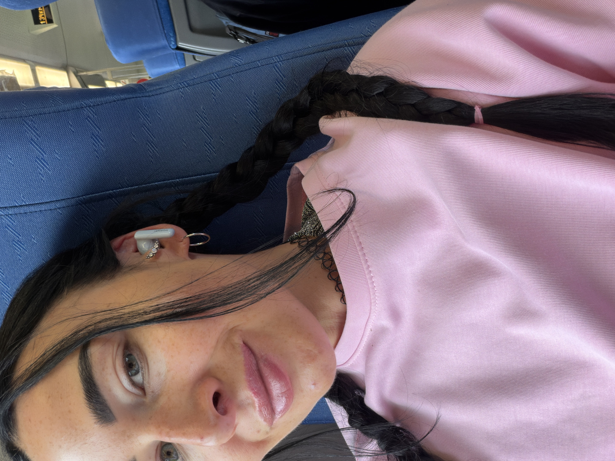 This media file is a close-up, portrait-style selfie of a young person with dark hair, wearing a pink sweatshirt and a black choker necklace. The person has light-colored eyes, thick eyebrows, and appears to be wearing lip gloss. They have a white earbud in their right ear and a hoop earring in their left. One braid of their dark hair is visible, resting on their shoulder. The background suggests an indoor setting with blue seating, possibly on a train or bus, given the blue fabric and a glimpse of a window with reflections. A sign with Cyrillic text is visible in the upper left corner of the image. The image is oriented sideways.