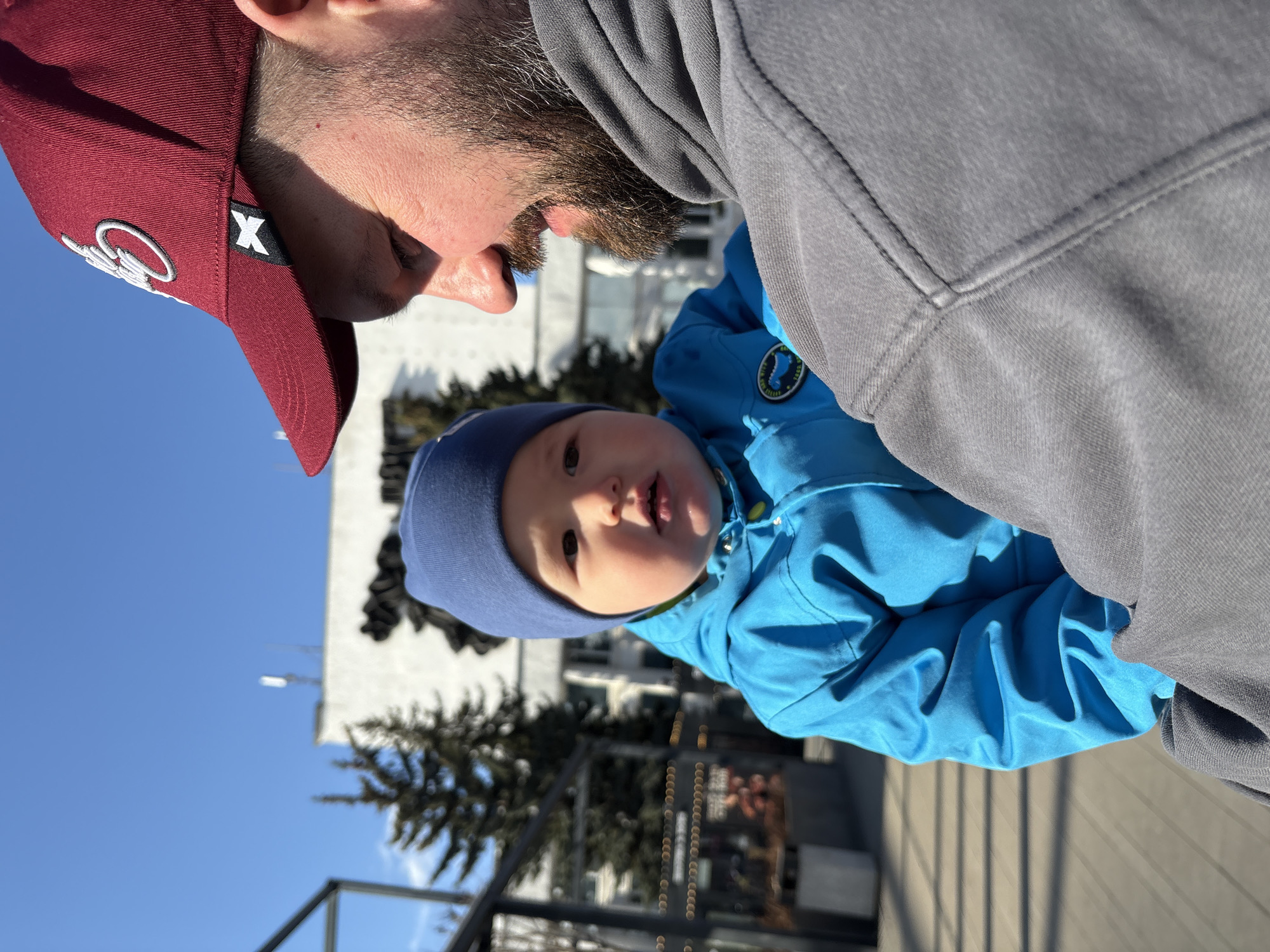 A close-up, partially obscured, ground-level shot depicts a man holding a baby outdoors. The man wears a maroon baseball cap with white embroidery on the front and a gray hooded sweatshirt. The baby is dressed in a blue snowsuit and a dark blue knit hat. The man and baby are positioned such that the baby faces the camera. The background includes a partially visible building, leafless trees, and a clear blue sky. The location is identified as Izhevsk, Russia.