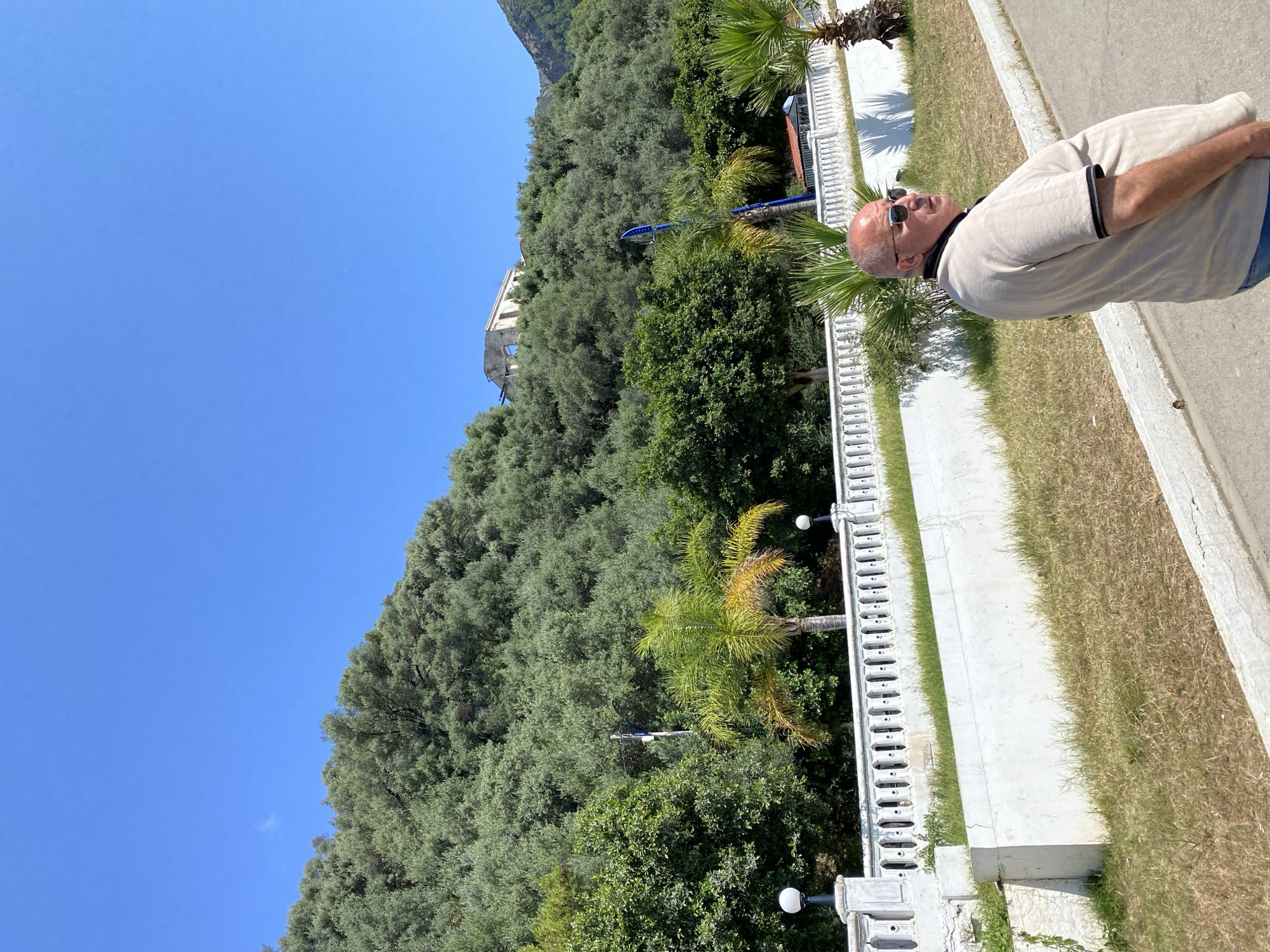 This full-shot image captures a sunny outdoor scene in what appears to be Bejaia, Algeria. The photograph is oriented vertically, with a man standing on the right side of the frame, a white balustrade and vegetation in the middle, and a dense, tree-covered hillside dominating the left.
In the foreground, a light-skinned man with a bald head and dark sunglasses stands on a paved path. He is wearing a light-colored polo shirt with dark trim on the collar and sleeves, and blue trousers. He faces left, seemingly observing the landscape. To his left, a white concrete balustrade with a series of decorative lamps runs horizontally across the midground. Behind the balustrade, there's a strip of green and dry grass, followed by lush green vegetation including several palm trees, one with yellowish fronds.
The background on the left features a steep hillside covered entirely by a dense forest of green trees, likely Mediterranean species such as olive trees. High on the hillside, a partially visible white building, possibly an old structure or ruins, peeks through the foliage. A small blue object, possibly a boat or a canopy, is also visible among the trees. The sky above is a clear, vibrant blue, without any clouds.
The image depicts a bright and clear day, suggesting strong sunlight and warm weather, consistent with a Mediterranean climate. No specific activity beyond standing and observing is taking place, and no text is visible. The scene evokes a sense of tranquil outdoor leisure.