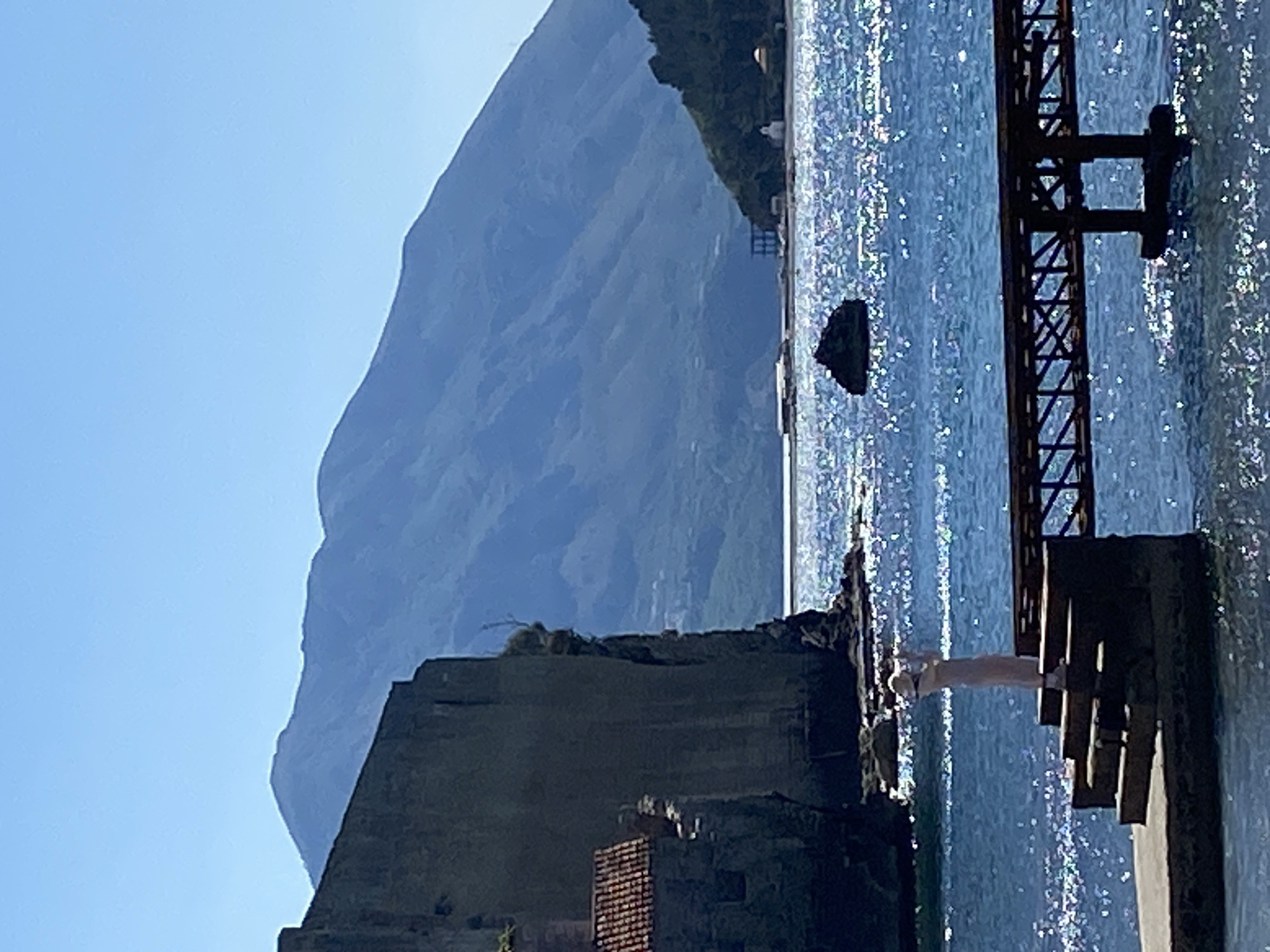 The image shows a view in Budva, Montenegro, featuring a body of water with reflections, a mountain in the background, and various structures. In the water, there's a prominent rock formation and a pier or dock structure with metal supports extending into the water. Part of a concrete or stone building is also visible in the foreground, partially obscuring the view. The sky is clear and blue. A person is partially visible in the water.