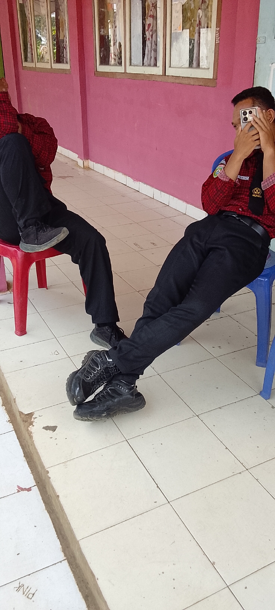 In Tanjung Setia, Indonesia, two individuals are seated on plastic chairs outside. The person on the left, wearing a red and black plaid shirt and black pants, is seated on a red chair with their left leg crossed over their right. They are wearing dark sneakers. The person on the right, also in a red and black plaid shirt and black pants, is seated on a blue chair. This individual is holding a smartphone up to their face, obscuring it. They are wearing black sneakers. The environment consists of a tiled floor and a pink wall with windows. The overall scene appears to be an outdoor waiting area or common space.