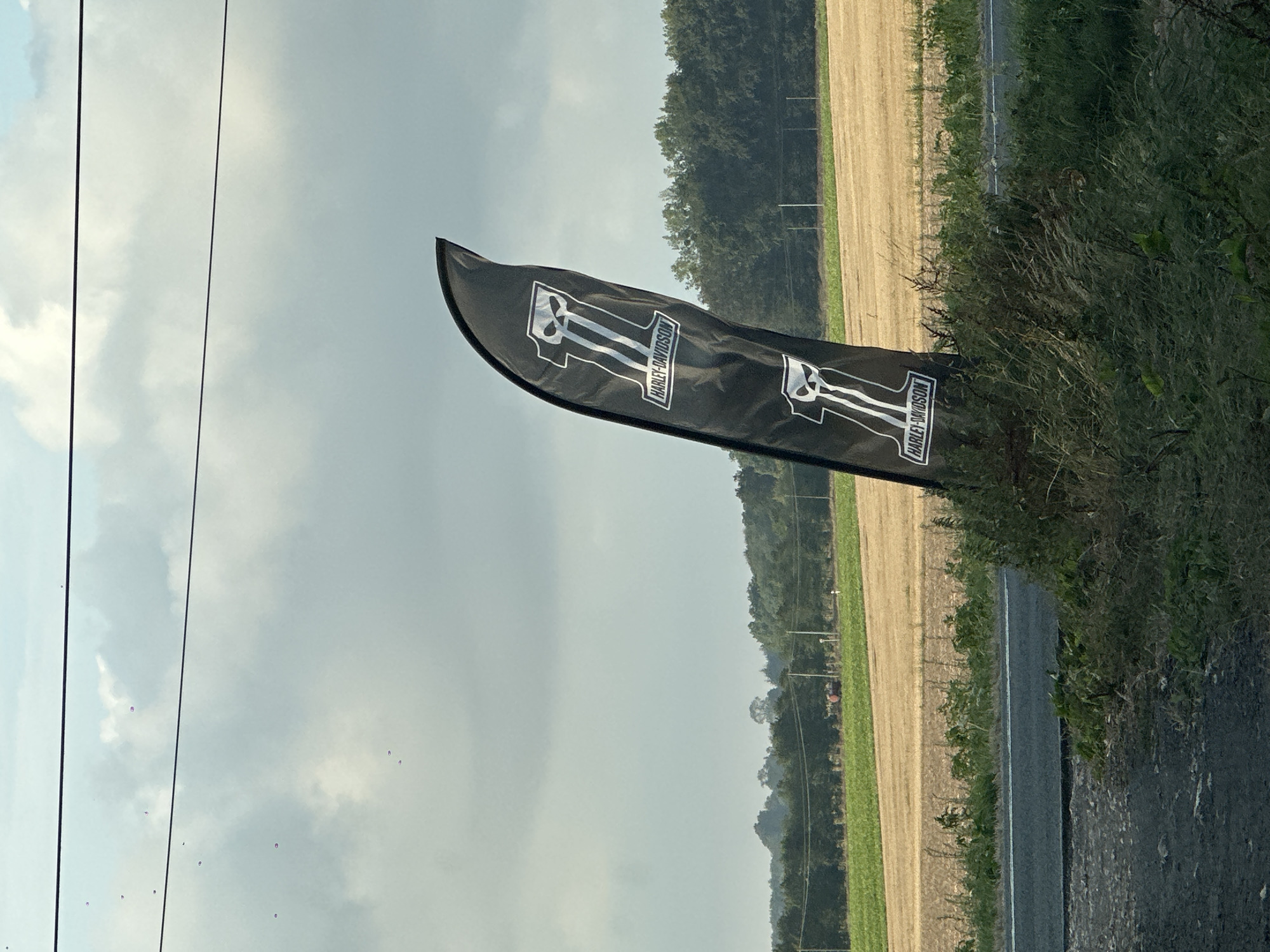 The image displays a black, feather-shaped flag with the Harley-Davidson logo printed in white. The logo features the Roman numeral "II" with a skull graphic at the top and the text "HARLEY-DAVIDSON" in a rectangular box below it. The flag is positioned in the foreground, leaning against a grassy embankment next to a road. In the background, there are fields of crops, trees, and a cloudy sky with power lines overhead. The scene appears to be an outdoor setting, possibly along a rural road. While the image is identified as being in Tôtes, France, no specific landmarks or identifying features of the city are visible in the photograph.