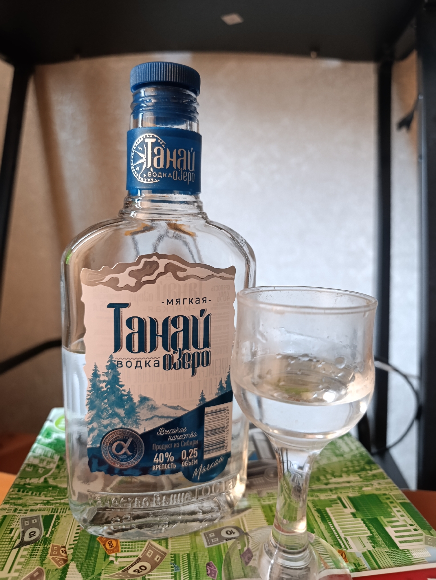 A bottle of "Tanay" vodka and a full shot glass are on a table. The bottle has a blue and white label with trees and mountains on it, and text in Russian that reads "Tanay" and "Vodka Ozero." The shot glass is clear and filled with a clear liquid. The table is covered with a green and white patterned cloth. The background is blurred and appears to be an indoor setting. No specific location in Barnaul, Russia can be determined from the image.