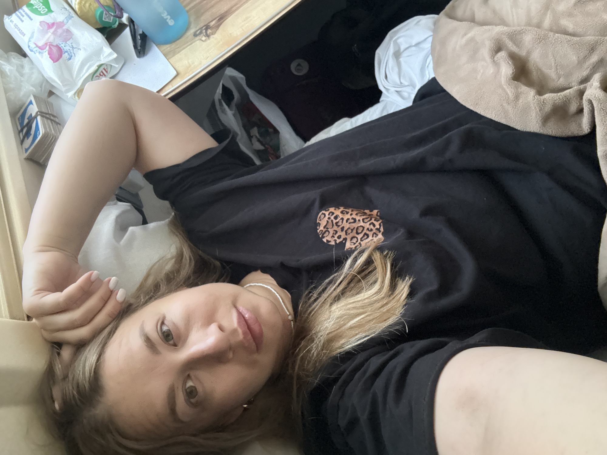 A young woman is depicted in a casual, head-and-shoulders selfie taken from a low angle while she is lying down indoors. She has light brown/blonde hair, light eyes, and fair skin, with a relaxed, slightly smiling expression. Her left arm is bent, with her hand supporting her head, showcasing light-colored nail polish. She is wearing a loose black t-shirt with a small leopard print design on the chest and a delicate silver necklace.
The setting appears to be a domestic space, likely a bedroom or living area. To her upper left, on a wooden surface, a collection of items is visible. These include a stack of small cards or tiles, bound together, with a prominent 'G' letter on one. Beside these are various personal items: a white plastic package featuring a pink flower graphic and partially visible Cyrillic text, including "абс" and "ОРГАНА"; another smaller yellow-green item; and a translucent blue water bottle with "eve" printed on it. A beige fleece blanket is draped over part of the bedding on the right side of the frame, near some darker fabrics.
The overall mood is casual and intimate, capturing a moment of rest or lounging. There are no strong indications of the time of day, but it appears to be well-lit indoors. The location context is Sergach, Russia, though no specific visual cues for this are present beyond the Russian text on the package.