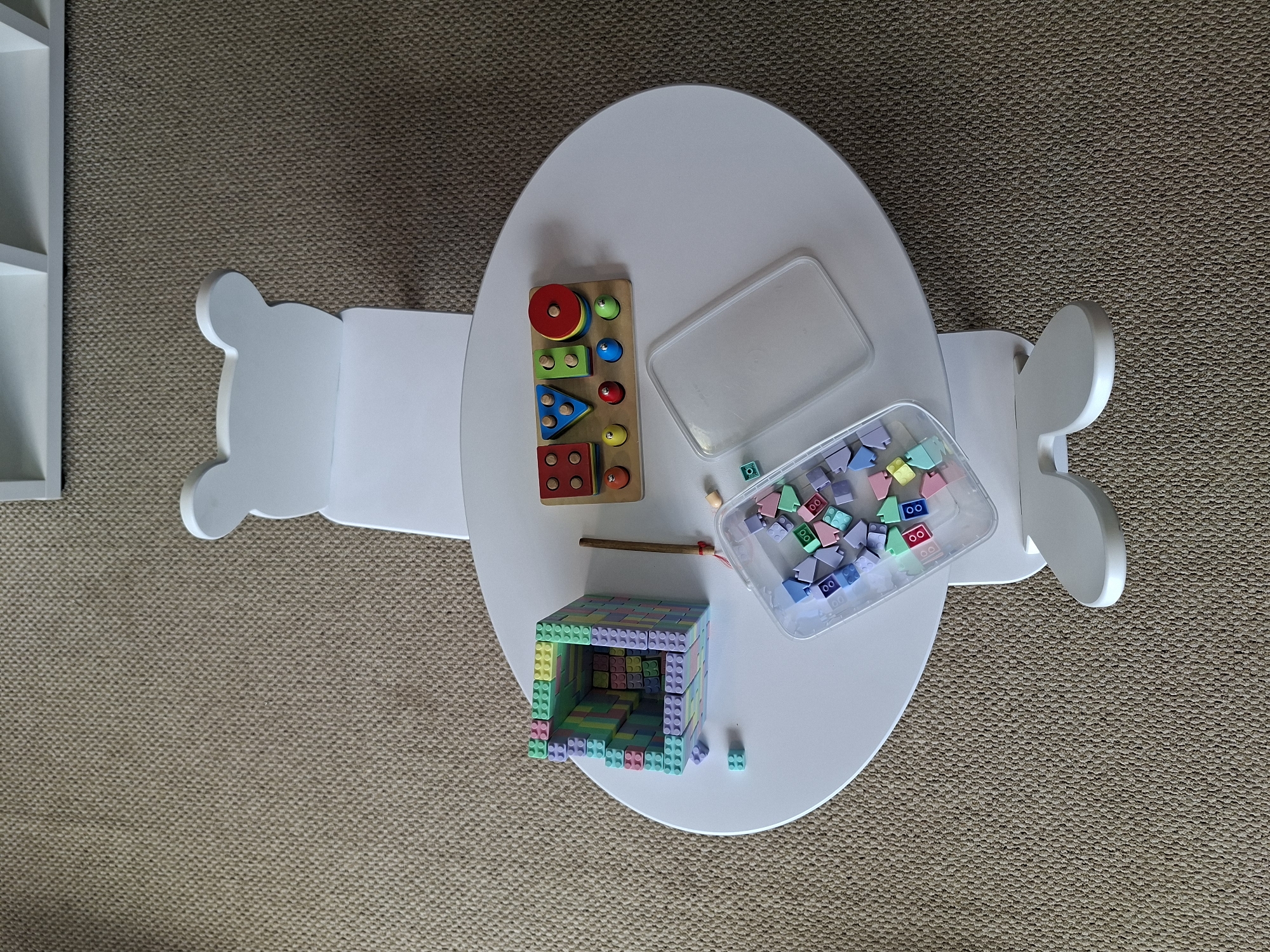 The image shows a top-down view of a white oval children's table with a bear-shaped backrest. On the table are several toys: a partially built structure of colorful interlocking blocks, a wooden shape-sorting toy with various geometric shapes and pegs, and a clear plastic container filled with loose interlocking blocks in pastel colors. A wooden stick with a red tassel is also present, lying next to the block structure. The table is situated on a textured, beige carpet. There are no people visible in the image. The scene appears to be a child's play area. The image does not contain any information that would allow for determination of its location in Izhevsk, Russia.