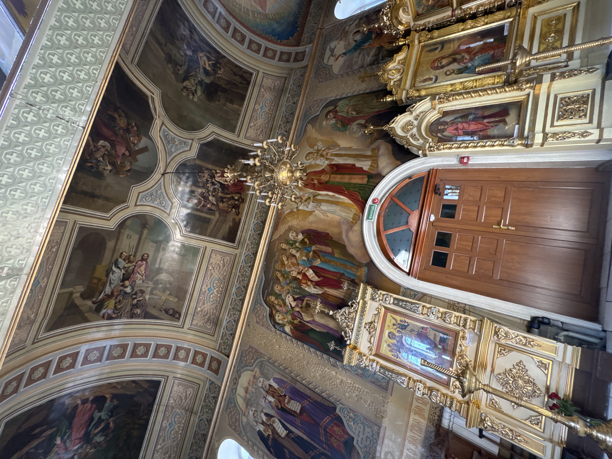 The image shows the interior of a richly decorated church, likely an Orthodox church given the iconography. The ceiling is adorned with large, detailed frescoes depicting religious scenes. A grand chandelier hangs from the center of the ceiling. The walls are also covered with murals and iconostasis, which are ornate screens covered with religious icons. Two large, gilded iconostases are prominently displayed, featuring framed religious paintings. A large wooden door with arched windows is visible in the center of the image, framed by the iconostases. In the foreground, to the left, a golden candlestick with red roses is placed. The overall impression is one of opulence and religious devotion.