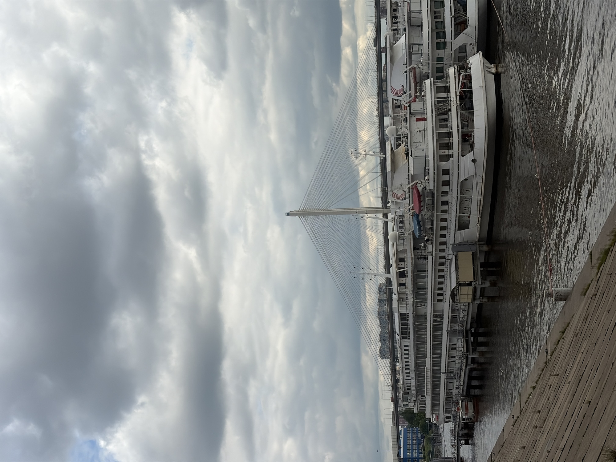 The image depicts a cityscape with a focus on maritime and architectural elements, possibly taken in Saint Petersburg, Russia. A large, multi-decked passenger ship is docked alongside a concrete embankment. The ship is white with numerous windows and balconies. A cable-stayed bridge with a tall, central pylon extends into the cloudy sky, its cables fanning out towards the river and surrounding structures. The sky is overcast with heavy clouds. The water appears to be calm. Along the embankment, there are signs of urban development, including buildings and structures. The overall scene suggests a riverfront area in a city with significant maritime traffic and modern infrastructure.