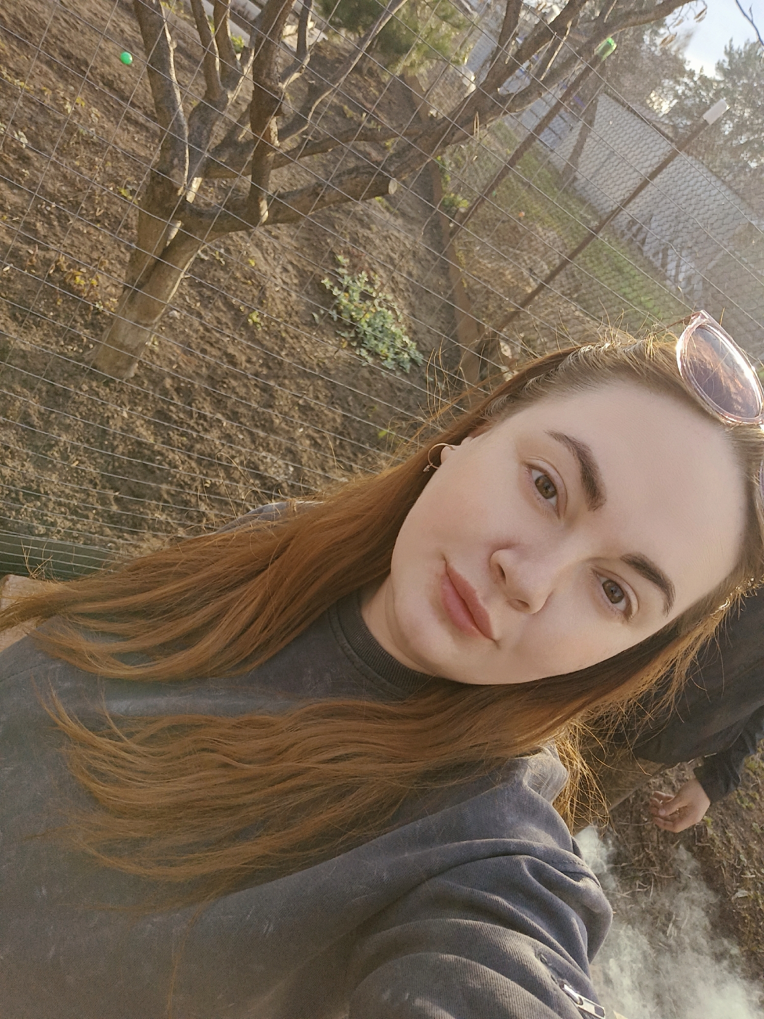 A woman with reddish-brown hair is seen in a selfie. She is wearing a grey sweatshirt and has sunglasses perched on her head. The background includes a tree and a wire fence, with a dirt-covered ground. White smoke is visible near the bottom of the image. This location is likely in Chaikovsky, Russia.