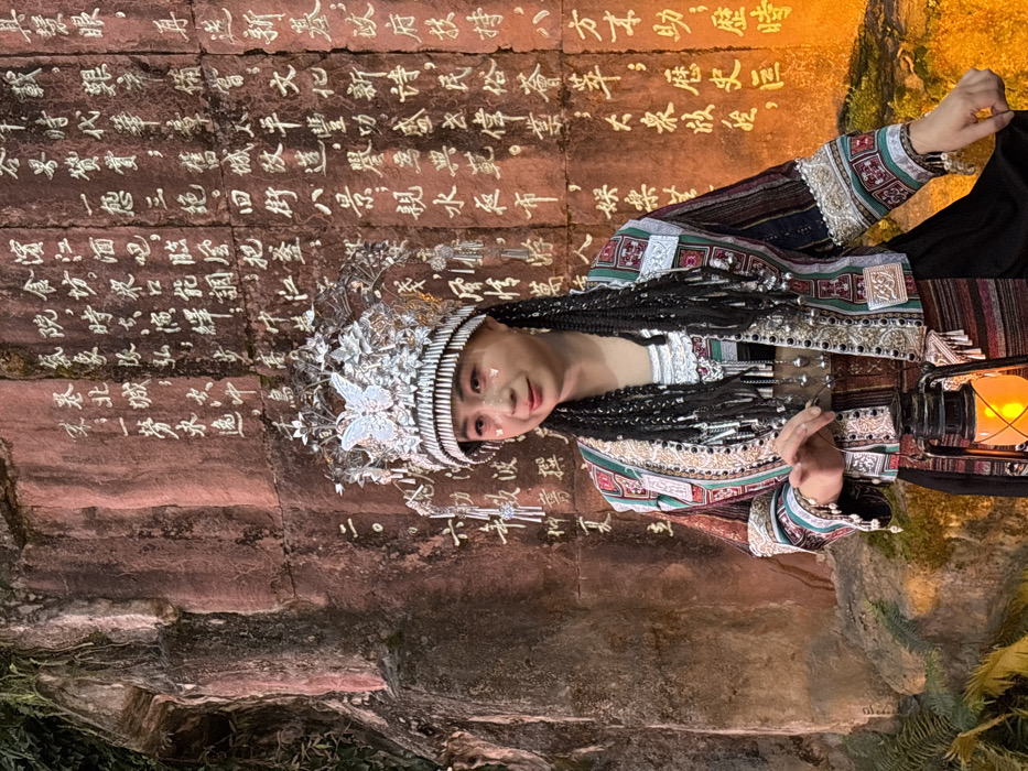 An adult female is depicted in a medium shot, standing in front of a reddish-brown rock wall in Yuzhong District, China. The wall is covered extensively with white Chinese characters and features some green foliage clinging to its left side. The woman faces slightly to the viewer's right, looking forward with a subtle smile. She wears an elaborate silver headdress, featuring a prominent central butterfly motif and intricate floral-like designs with dangling silver ornaments. Her face is adorned with makeup, including white shimmering accents on her cheekbones and dark red lipstick. Her long, dark hair is styled into numerous thin braids, each tip decorated with small silver beads. She is dressed in a multi-colored, richly embroidered cropped jacket with patterns in green, white, red, brown, and black, accented with silver-colored trim and embellishments. Beneath the jacket, a silver-colored neckpiece or necklace with multiple dangling silver bells is visible over a dark garment. A striped, woven sash in earthy tones encircles her waist. In her left hand, she holds an antique-style lantern emitting a warm, orange glow. The scene is illuminated by warm, directional light, highlighting the subject and the carved wall.