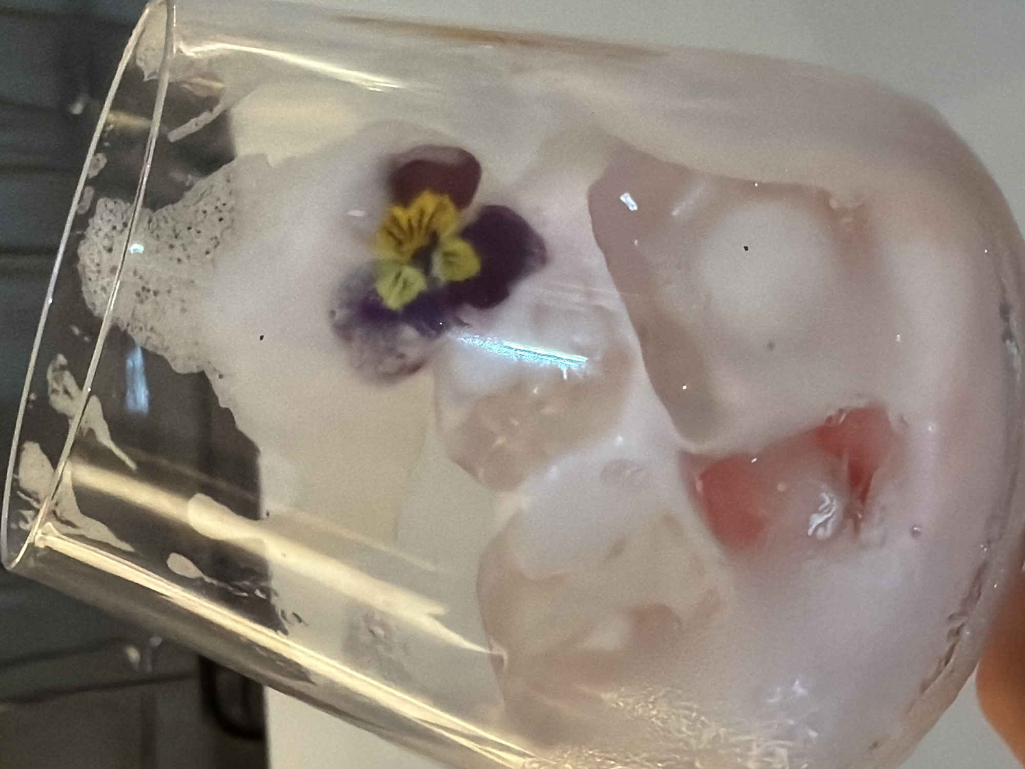 A close-up shot of a cocktail in a clear glass. The drink contains ice, foam, and a small purple and yellow flower. A piece of fruit appears to be encased in the foam near the bottom of the glass. The shot is taken indoors, likely in a bar or restaurant. The glass is tilted slightly. The background is indistinct. No people are visible. No location in Bishkek City, Kyrgyzstan can be identified.