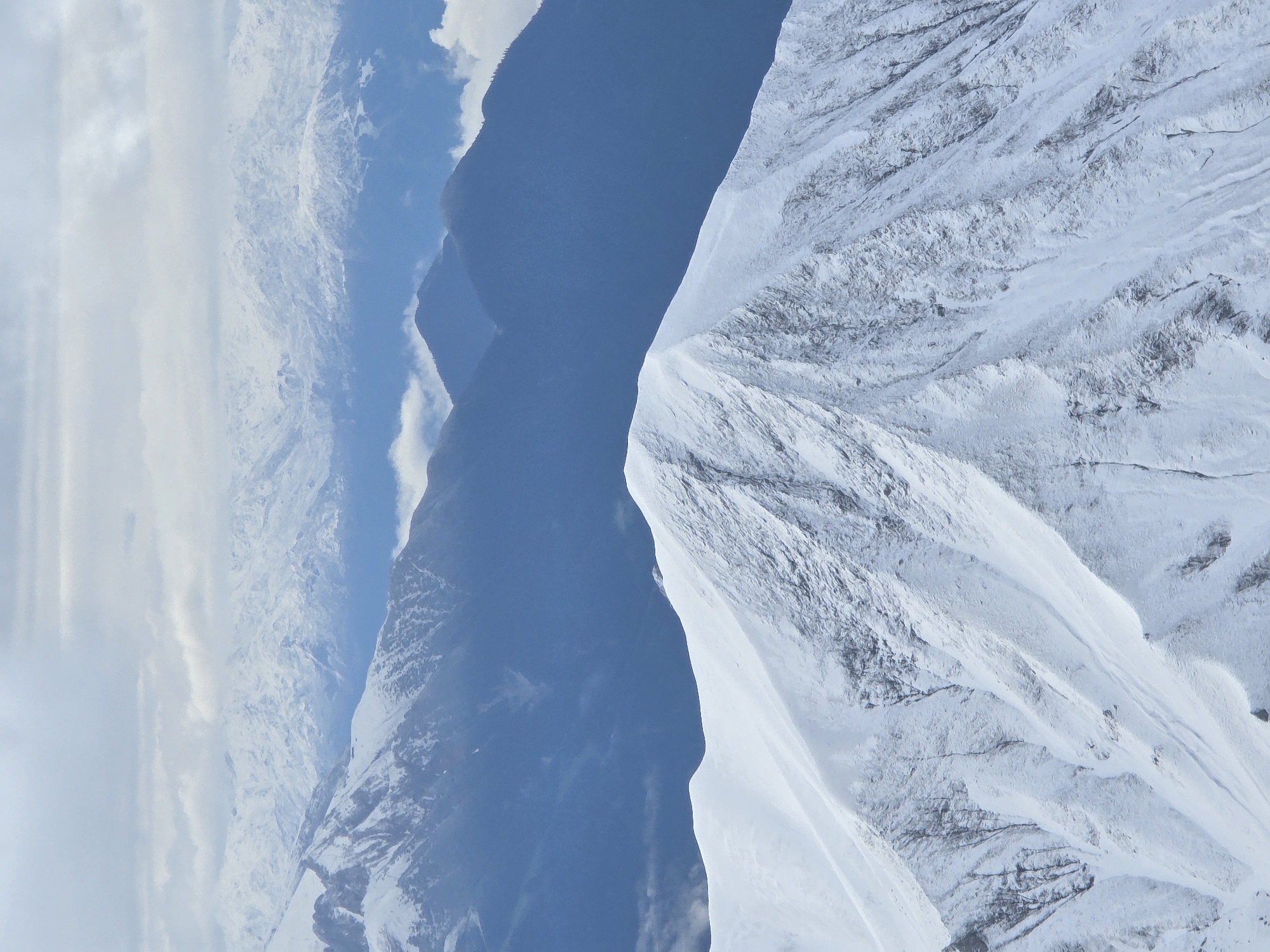 This is a vertical, eye-level, close-up shot of snow-covered mountains. The foreground is dominated by a steep, snow-covered mountain peak with visible rocky textures beneath the snow. The middle ground features a series of blue-toned mountain ranges receding into the distance, some with lighter snow caps and others appearing darker and more shadowed. The background is filled with layers of distant snow-capped mountains, fading into a hazy, overcast sky with scattered clouds. The scene is natural and depicts a mountainous landscape, likely taken from a high vantage point. There are no people or man-made objects visible in this image, suggesting a remote natural environment. The image does not contain any visual information that places it specifically in Garmisch-Partenkirchen, Germany.