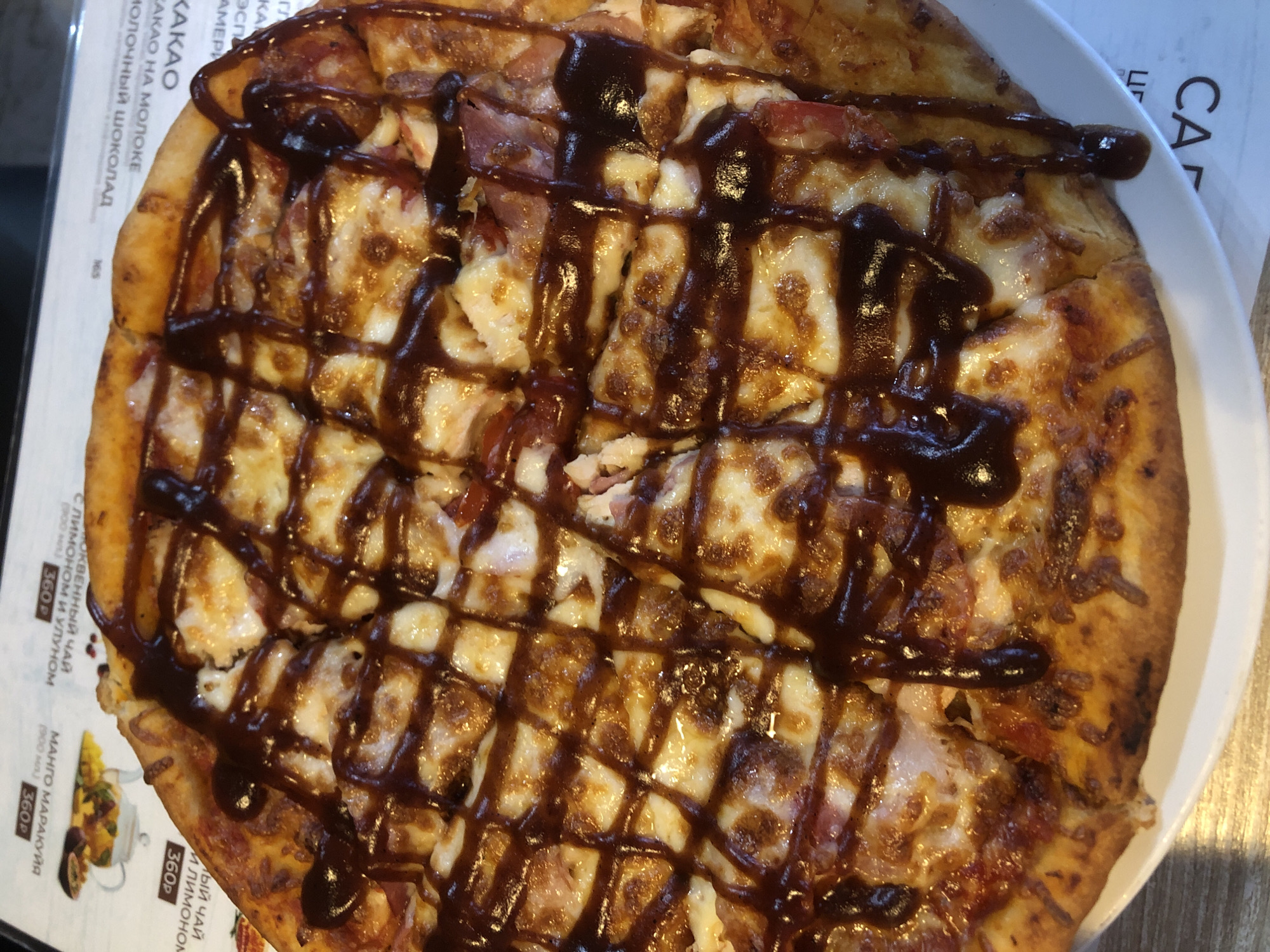 The image displays an overhead close-up of a circular pizza, freshly prepared and served on a plain white plate. The pizza has a golden-brown crust and is topped with melted, bubbly cheese, interspersed with pinkish-brown meat pieces, likely ham or bacon. A thick, dark brown barbecue sauce is drizzled in a prominent grid pattern across the entire surface of the pizza.
The plate rests upon a white menu or placemat featuring Cyrillic text and small graphical elements. Visible text includes "МАНГО МАРАКУЙЯ" (Mango Passion Fruit) priced at "360P" (360 Rubles), and part of "МОЛОЧНЫЙ ШОКОЛАД" (Milk Chocolate). A partial brand name "САГ" is also visible at the bottom right. The underlying surface is a light-colored wooden table. The scene depicts a dining setting within a cafe or restaurant located in Chaikovsky, Russia.