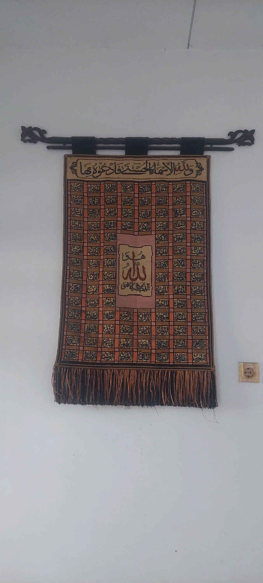 An Islamic tapestry featuring Arabic calligraphy is hung on a white wall. The tapestry is suspended from a dark, ornate metal rod. The fabric itself is rectangular with a fringe along the bottom. The background of the tapestry is a checkered pattern of orange and brown squares, each containing a section of Arabic script. In the center of the tapestry, a light pink rectangular panel displays a prominent calligraphic rendering of "Allah" in the upper portion, and another phrase in Arabic below it. To the right of the tapestry, a light-colored electrical outlet is visible on the wall. The image is taken from a frontal perspective, with the tapestry centrally placed.