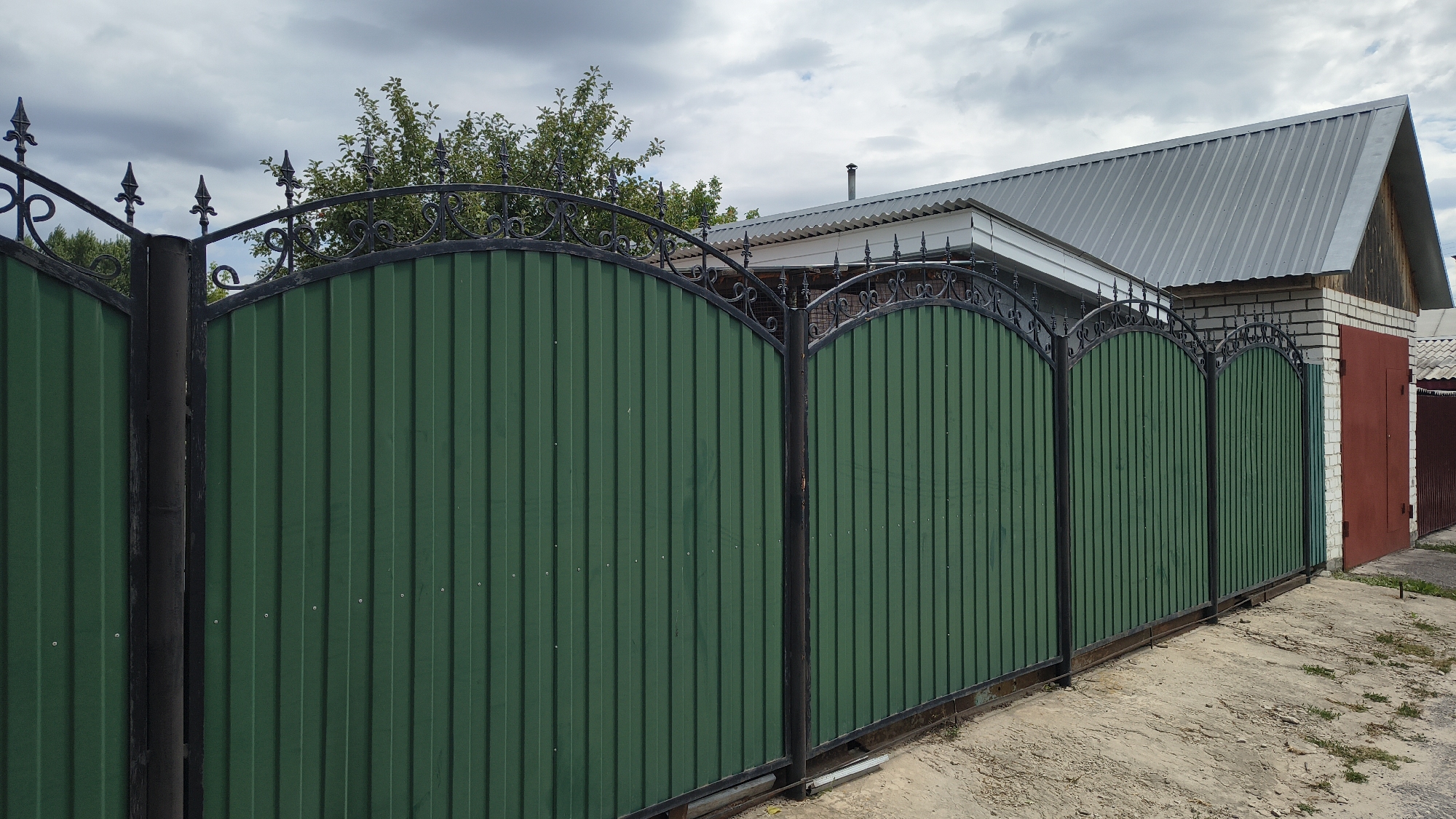 A dark green corrugated metal fence with decorative wrought iron accents runs along the right side of the frame. The fence is segmented into several arches, with a black metal post separating each section. Behind the fence, a building with a corrugated metal roof is visible, as well as a red garage door. Trees are seen behind the fence on the left side of the frame. The ground in front of the fence appears to be a dirt and gravel path. The sky above is overcast with grey clouds. The location is the city of Mikhaylovka, Russia.