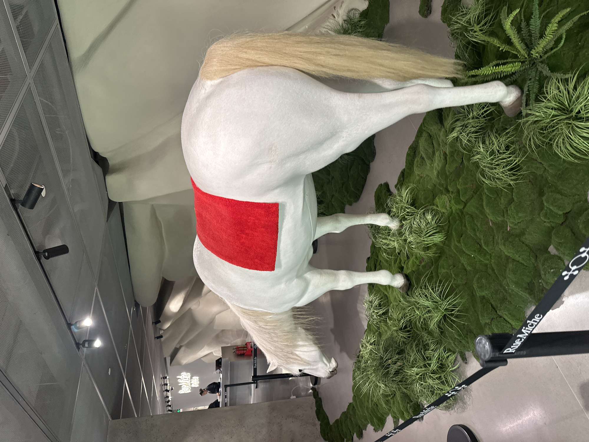 The image depicts a white horse statue with a red blanket on its back, positioned on a bed of artificial green moss and plants. A black rope labeled "Rue Miche" surrounds the display. The location is an indoor space, likely a retail or exhibition area, with a modern design featuring a grey concrete floor and ceiling grid. In the background, there are people and displays suggesting a commercial environment. The exact location in Thủ Đức, Vietnam is not discernible from the image.