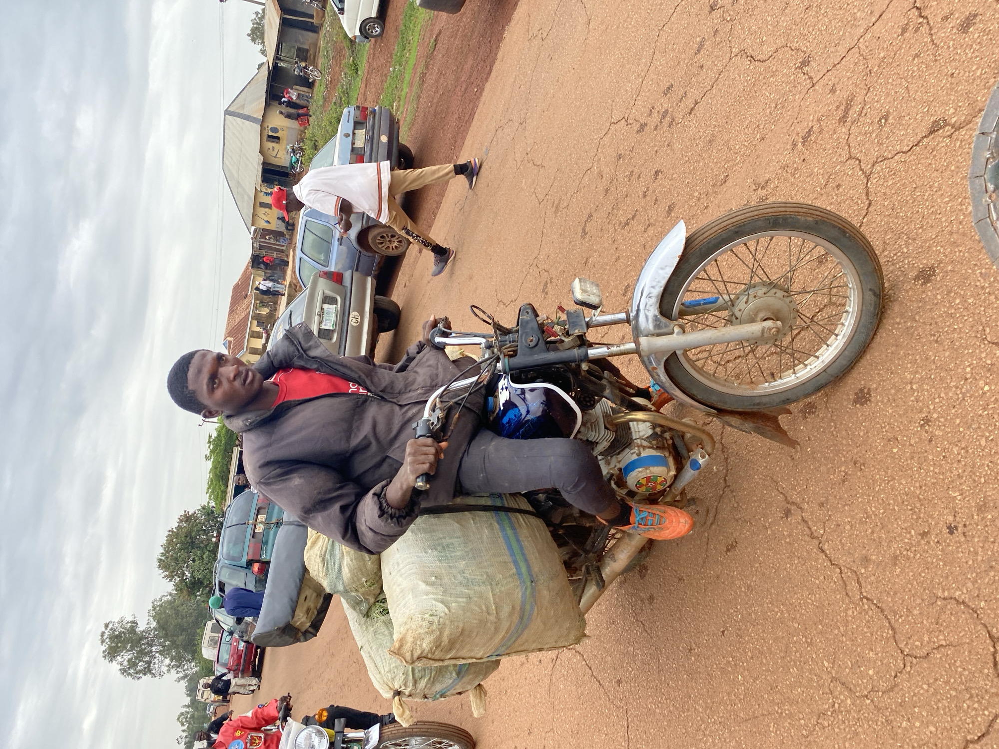 A man in a red shirt and black jacket sits on a motorcycle loaded with large sacks on a street in Durimi, Nigeria. He wears grey pants and bright orange shoes. In the background, cars are parked on the side of the road, and a man in a white shirt and khaki pants walks nearby. Buildings are visible in the background under a cloudy sky. The road appears to be cracked and reddish-brown in color.