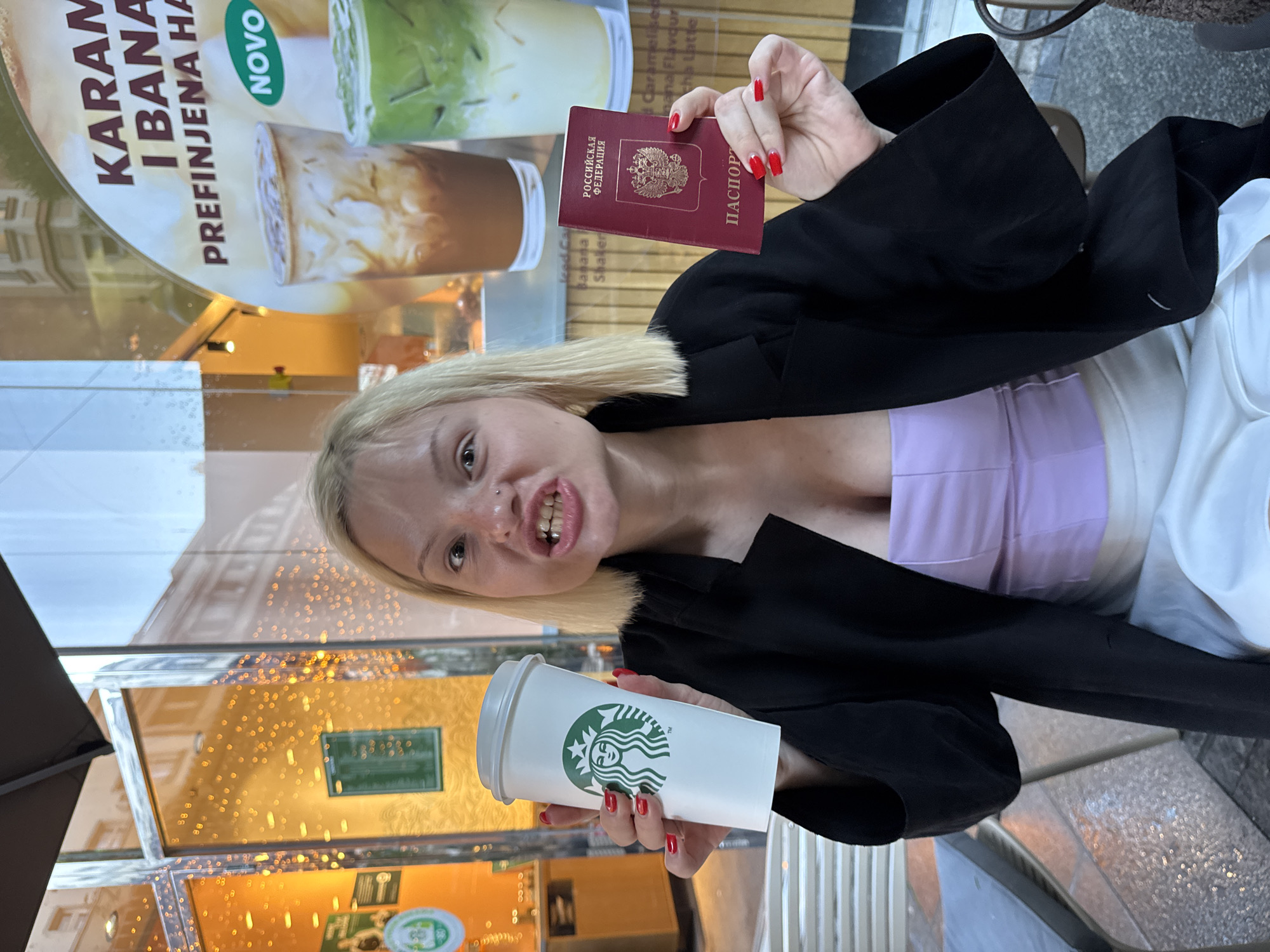A fair-skinned, blonde-haired woman with red nail polish holds a white Starbucks coffee cup with a green logo in her left hand and a burgundy-colored Russian passport in her right hand. She is wearing a black blazer over a lavender top. Behind her is a window display advertising "Karamel I Banana Prefinjena Ha" from Starbucks in Belgrade, Serbia. The outdoor scene includes glittering lights and a portion of a cafe umbrella. The woman appears to be posing for the photo.