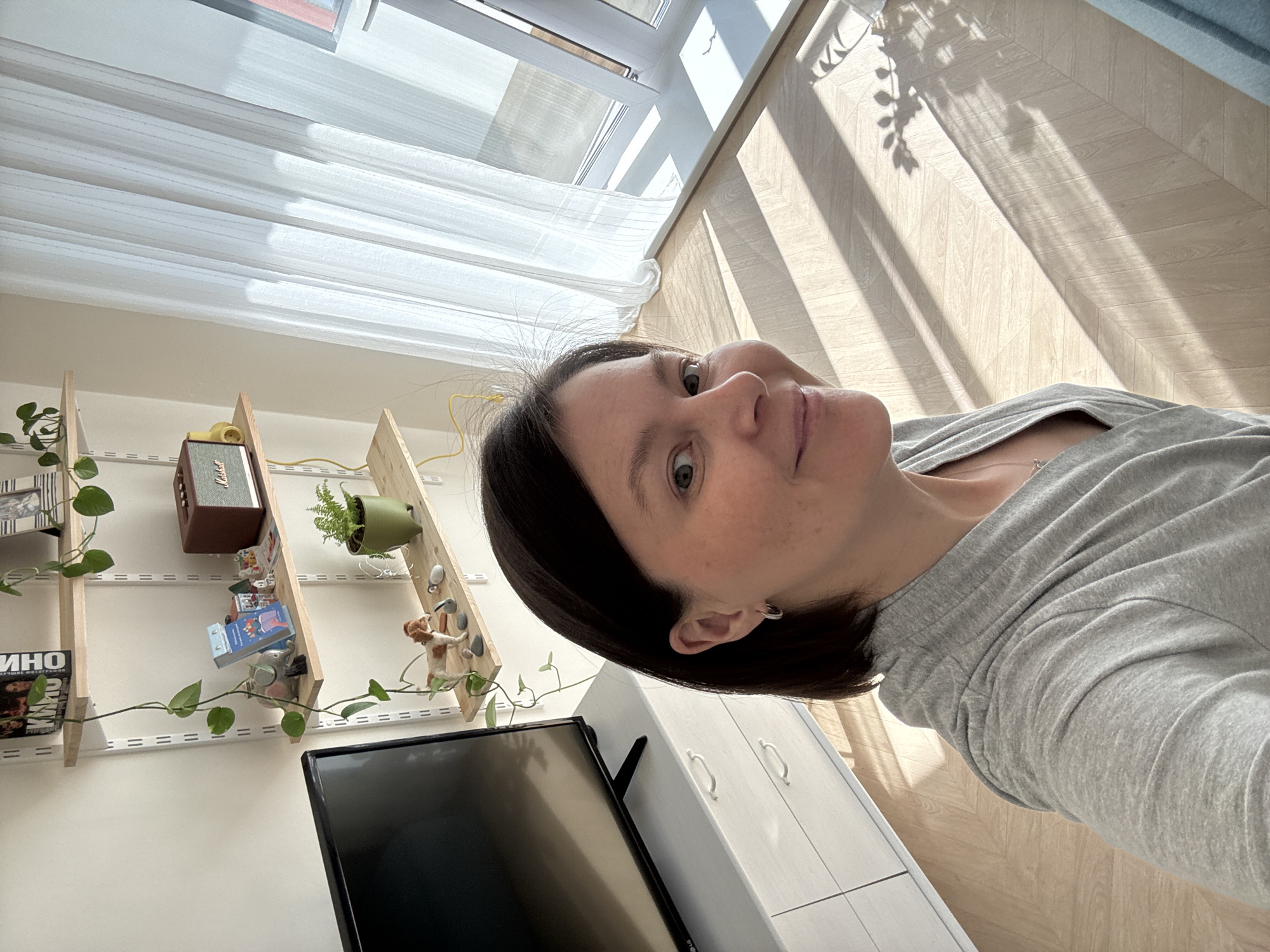 A selfie of a woman in a gray shirt taken indoors. In the background is a window with a white curtain, a wooden floor with shadows from foliage, a bookshelf with a speaker, books, a plant, and small figurines. A television and a white cabinet are also visible. The location is identified as Izhevsk, Russia.