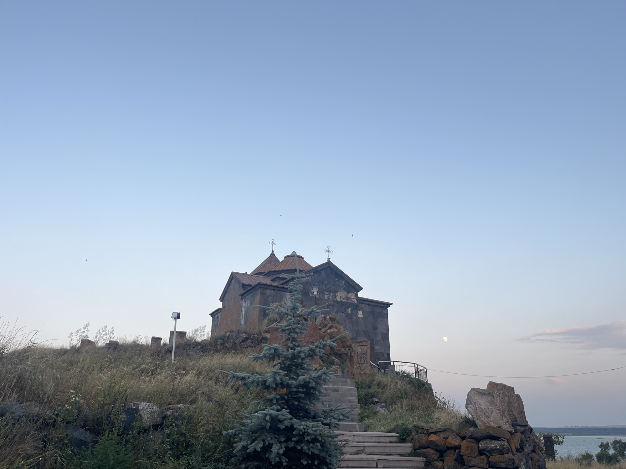 This is a landscape photograph of a church situated on a hill. The sky is clear and blue, with a gradient of lighter blue towards the horizon. In the foreground, a few small trees and dry grass cover the uneven terrain. A set of stone steps leads up to the church, which is made of dark stone with a reddish roof. The church has two domes, each topped with a cross. To the right of the church, a stone wall is visible, constructed with rough-hewn rocks. In the background, there is a body of water, likely a lake, with a distant shoreline. The moon is visible in the sky, high and to the right of the church. The overall atmosphere is peaceful and serene. The church in the image is likely the Sevanavank monastery in Hayravanq, Armenia, based on its distinctive architecture and location on a hill overlooking a body of water.