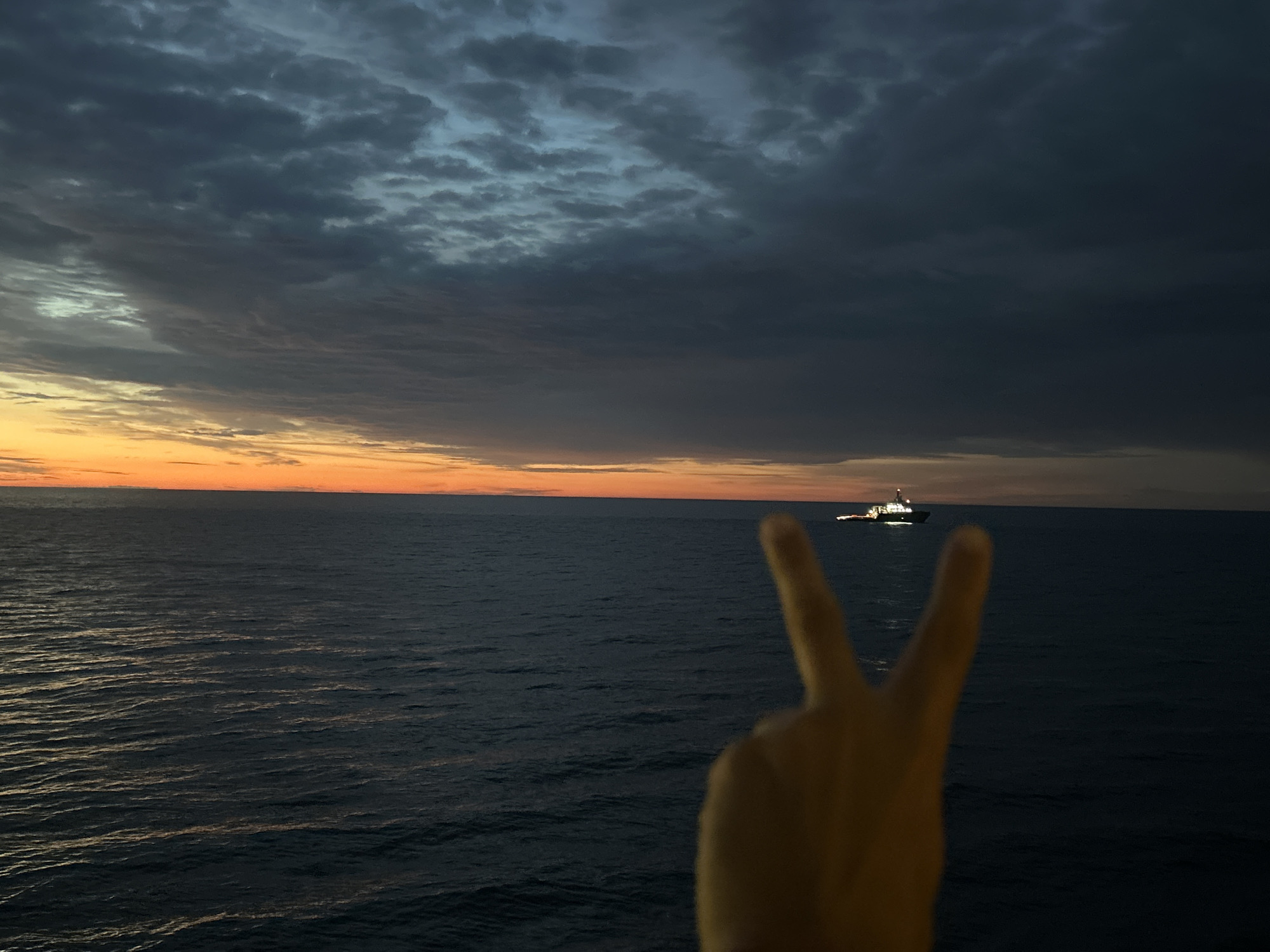 The image shows a hand making a peace sign in the foreground, with a body of water and a ship on the horizon in the background. The sky is filled with dramatic clouds, with a sliver of orange and yellow visible at the horizon, indicating either sunrise or sunset. The water is dark and appears choppy. The ship is a small vessel with lights on, suggesting it is either a working boat or a pleasure craft at sea. The overall mood of the image is serene and slightly mysterious. The location is Unknown, Unknown.