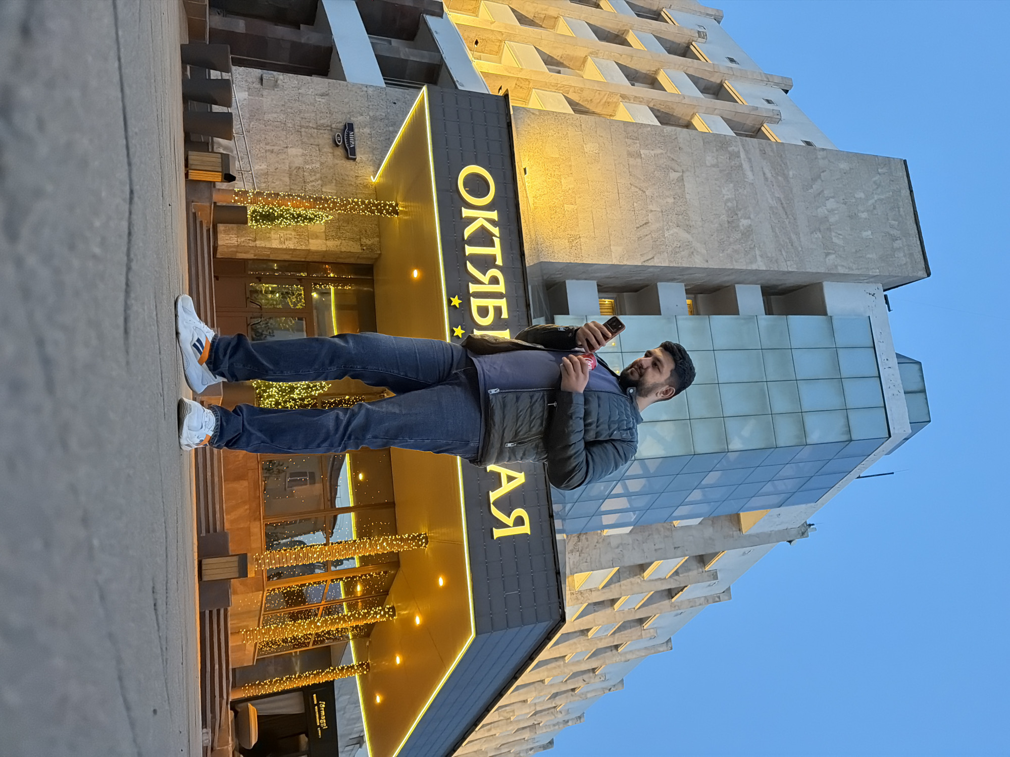 A man stands on the sidewalk, holding a phone, outside the "Oktyabrskaya" hotel in Krasnoyarsk, Russia. The hotel's entrance is brightly lit with golden lights. The sky is a pale blue, indicating twilight. The building itself has a mix of stone and glass architecture.