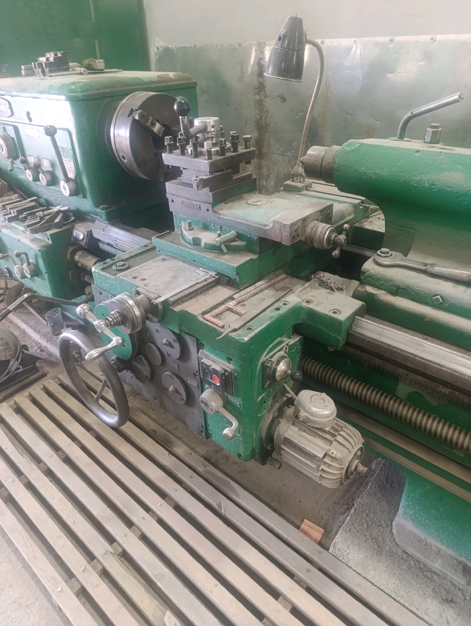 The image displays a large, green industrial lathe machine in a workshop setting. The machine is dusty and appears to be in a state of disuse or ongoing maintenance. Key components visible include the chuck, tool post, various levers and handwheels for operation, and a motor. The workshop floor is concrete, with a wooden slatted platform in the foreground. The background features a metal wall and a gooseneck lamp. The scene is static, with no people or ongoing activity depicted. The location is identified as Navoiy City, Uzbekistan.