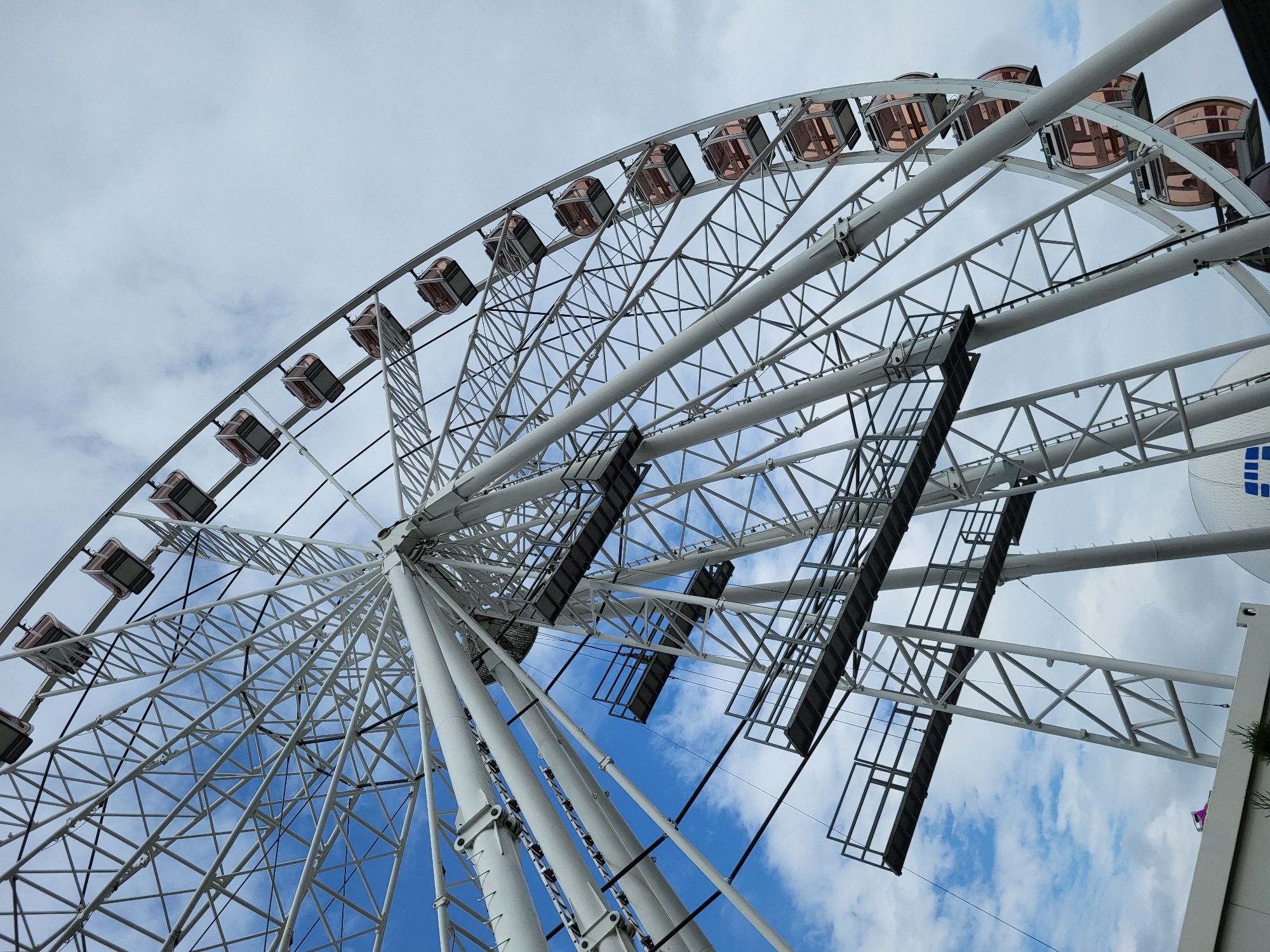 A low-angle shot captures a large, white Ferris wheel against a partially cloudy sky. The intricate metal framework of the Ferris wheel, composed of countless spokes and supports, dominates the composition. Numerous rectangular gondolas or cabins, featuring what appear to be rose-gold or bronze-tinted windows, are evenly spaced around the wheel's rim.
The sky transitions from a lighter, overcast grey-blue at the top left to a clearer, brighter blue with fluffy white clouds towards the bottom right. Two prominent dark grey, rectangular platforms or grates are visible on the inner structure of the Ferris wheel, possibly serving as maintenance platforms or mounts for lighting equipment.
The image is taken during daytime, indicated by the bright, diffused light. There are no people visible in the cabins or around the base of the structure. On the far right edge of the frame, a segment of a solid white structure with an indistinct blue and white geometric logo or pattern is visible, along with a small amount of green foliage. No specific location or legible text is discernible.