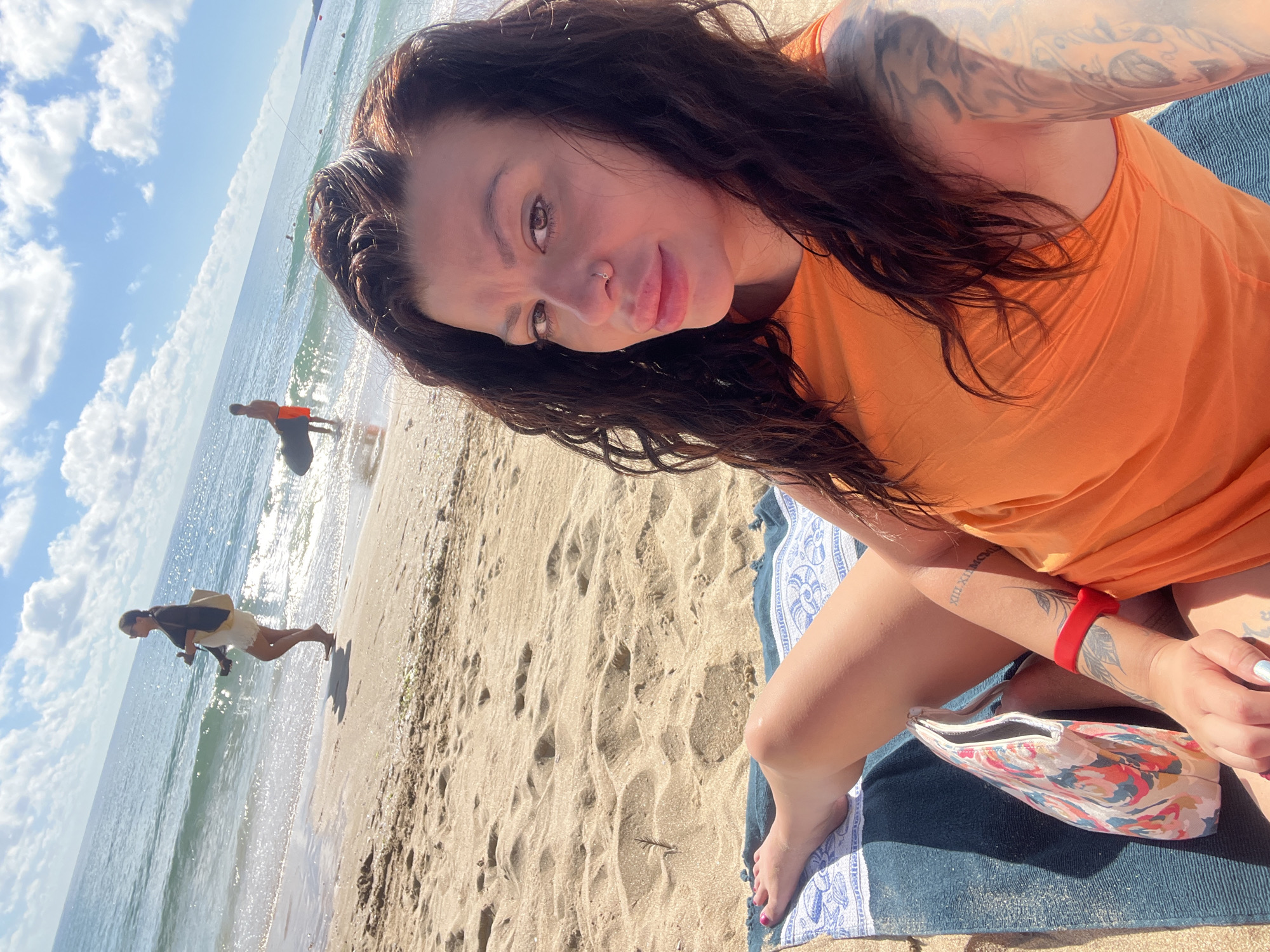 A woman is taking a selfie on a sandy beach in Sozopol, Bulgaria. She is lying on a towel and wearing an orange top. Her left leg is bent at the knee, and her left foot, with pink nail polish, is visible. A floral makeup bag is next to her. Her right arm has tattoos and a red bracelet. In the background, the sea is visible with waves. Several people are in the water and on the shore. One person is walking along the water's edge, and another is standing in the shallow water holding a surfboard. The sky is blue with scattered clouds. The sand is light brown and marked with footprints.