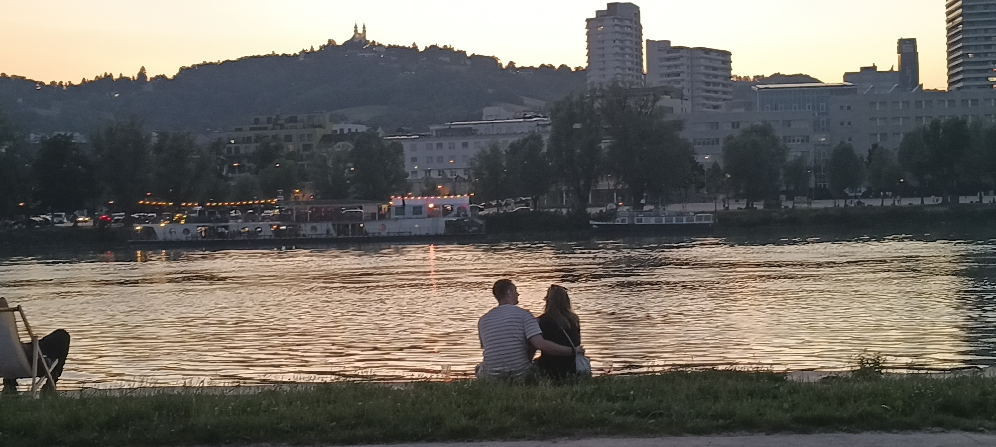 The image shows a couple sitting on the grassy bank of the Danube River in Linz, Austria, at sunset. The river's surface reflects the warm colors of the sky. In the background, across the river, is the cityscape of Linz. Visible elements include a hill with a prominent church or castle-like structure on its summit, various buildings of different architectural styles including apartment blocks and what appears to be office buildings, trees lining the riverbank, and several boats, one of which is a passenger boat with lights on. The couple is viewed from behind, with the man's arm around the woman. The scene is tranquil and romantic, capturing a moment of shared peace and contemplation of the cityscape as day transitions to evening.