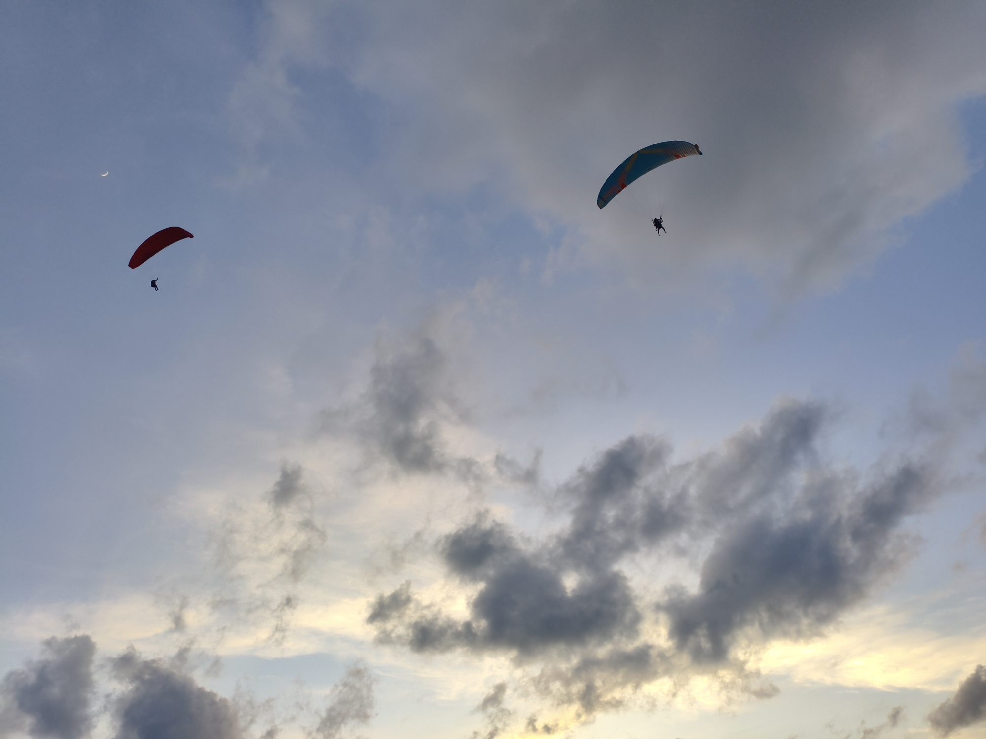 Two paragliders are visible in the sky over Rawai, Thailand. The paraglider on the left is red and in the upper left quadrant of the image. The paraglider on the right is multicolored, with stripes of blue, orange, and white, and is in the upper right quadrant of the image. Both paragliders have a person suspended beneath them, controlling the canopy. The sky is partly cloudy, with scattered gray clouds interspersed with patches of blue sky. A sliver of the moon is visible in the upper left corner. The lighting suggests it may be late afternoon or early morning, with some warmer tones from the setting or rising sun visible at the horizon, peeking through the clouds.